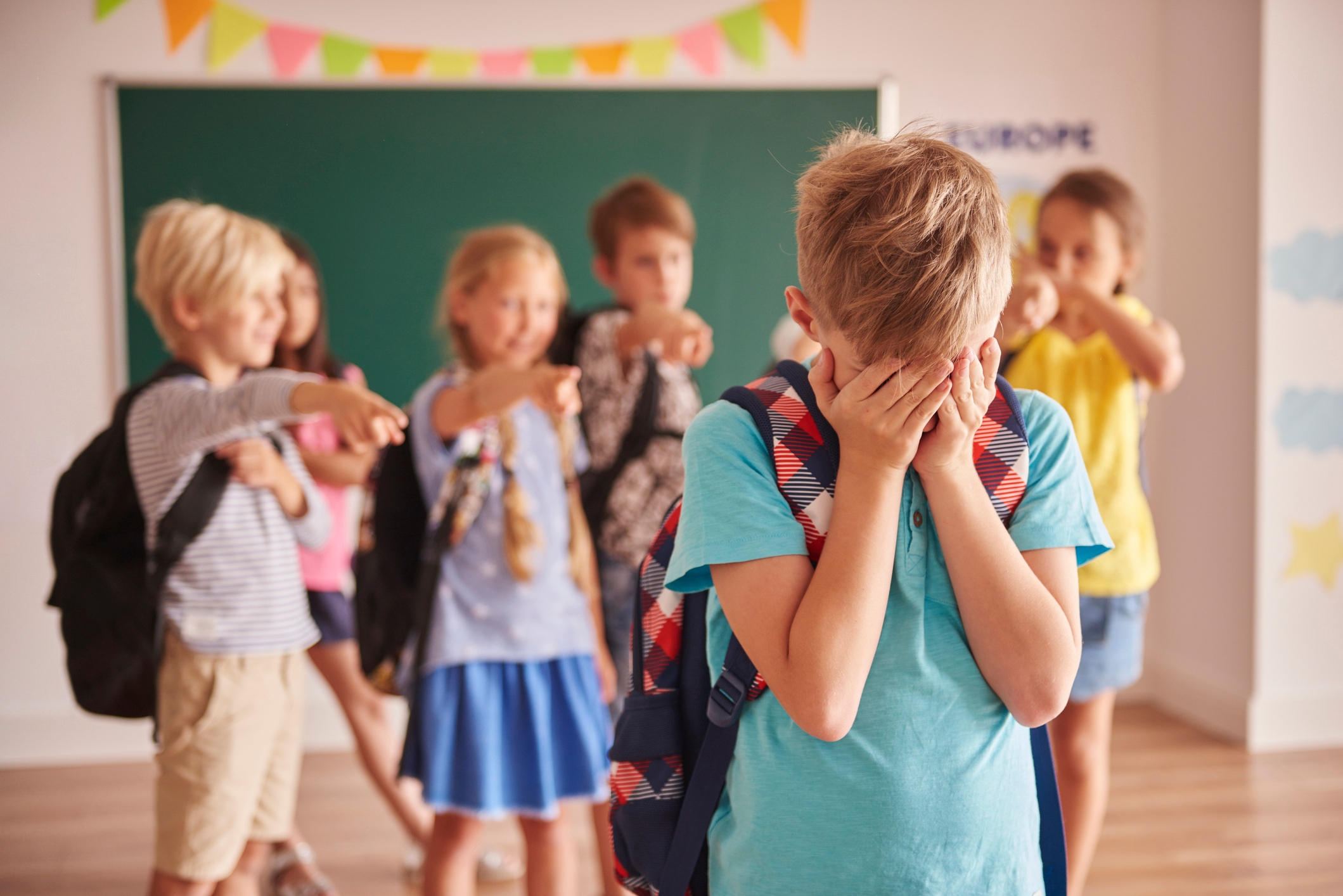 Child with backpack covers face, while peers point and laugh in classroom, illustrating school bullying theme