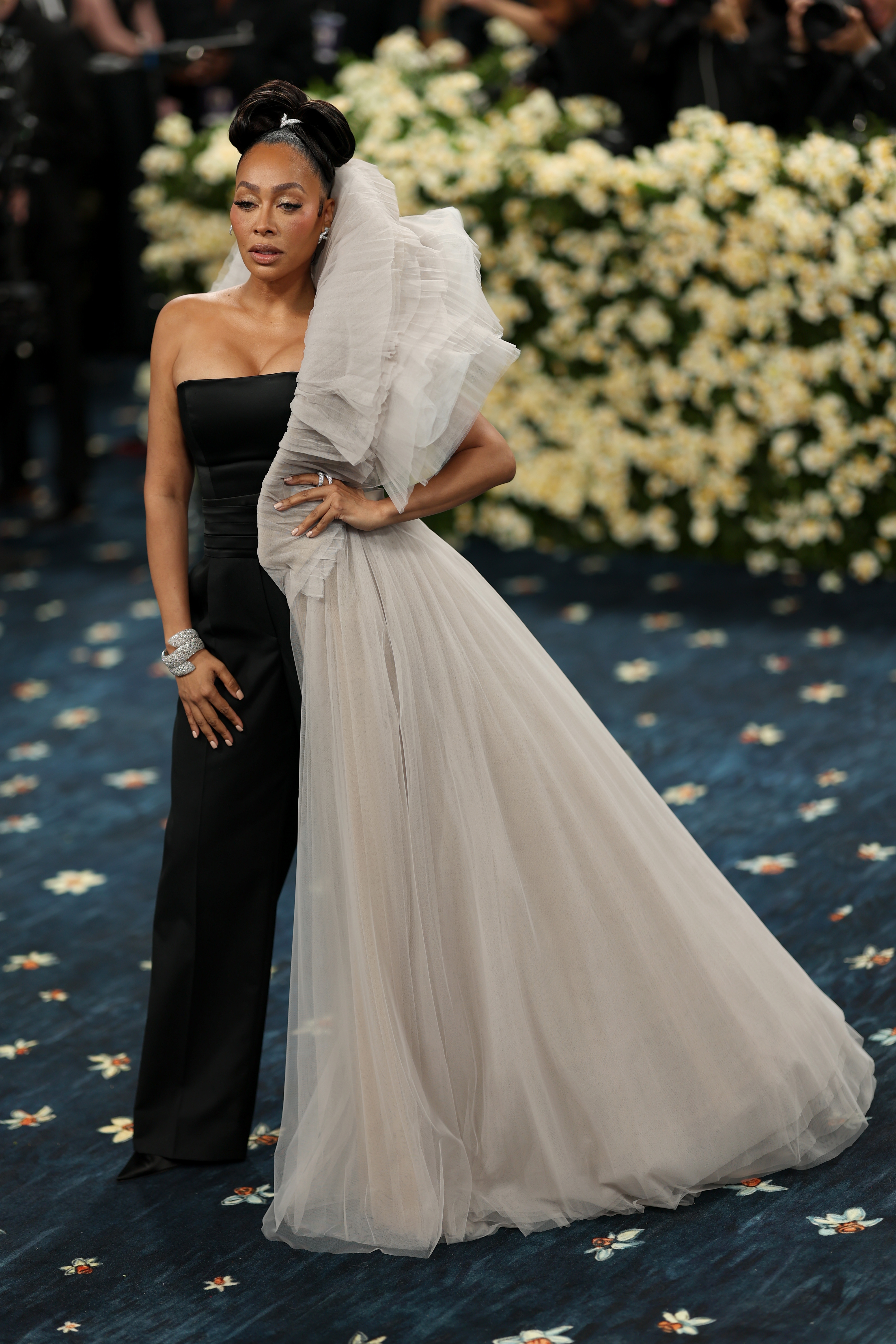 Woman in elegant black-and-white gown with tulle detailing poses on a floral-patterned red carpet at a celebrity event