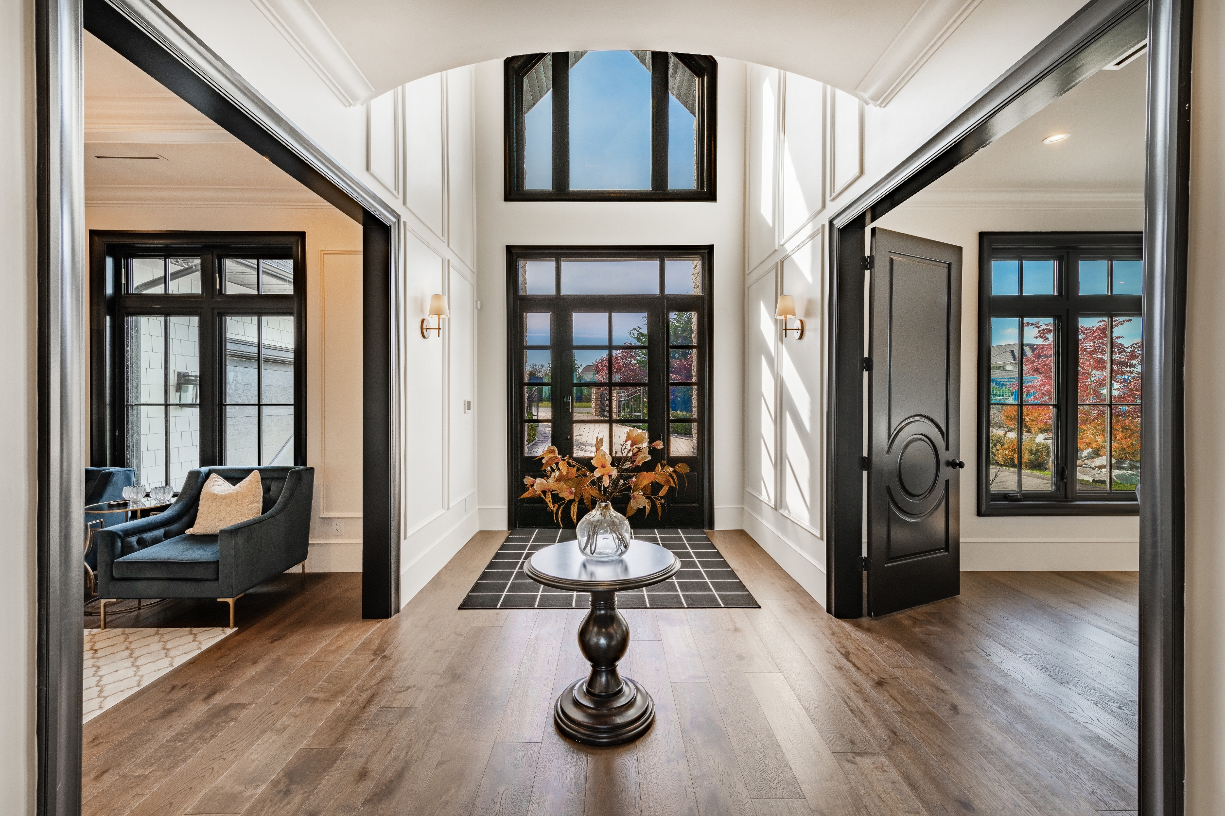 Spacious, elegant entryway with wooden floors, high ceilings, and large windows. A small table with a glass vase and dried leaves is centered in the room