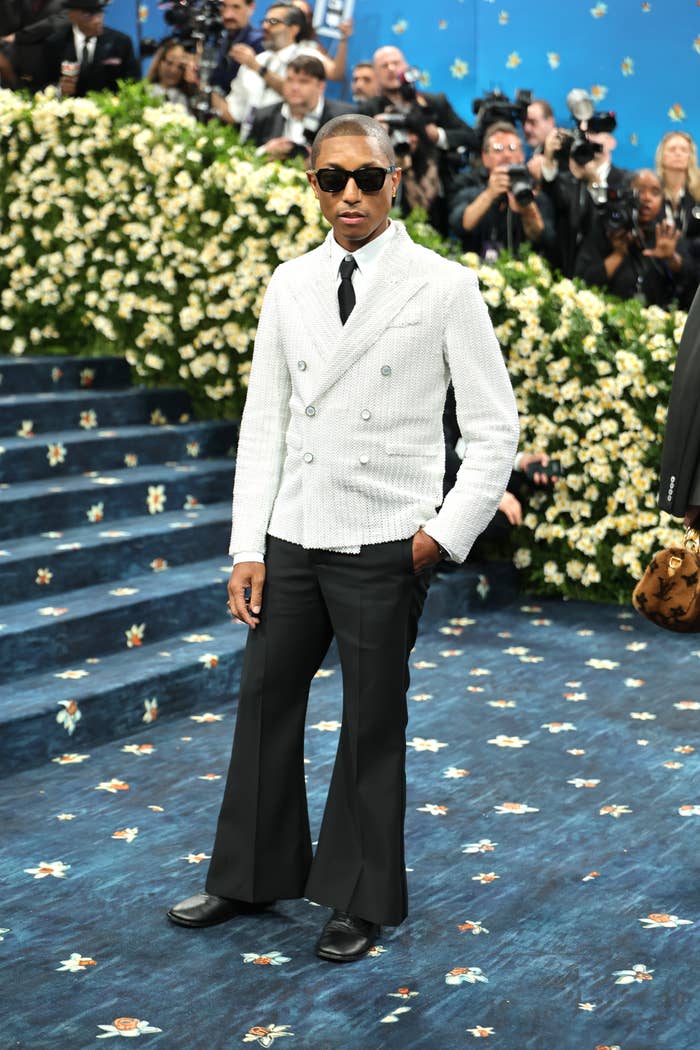 Person in a double-breasted jacket with wide-leg pants and sunglasses on a red carpet event with floral backdrop