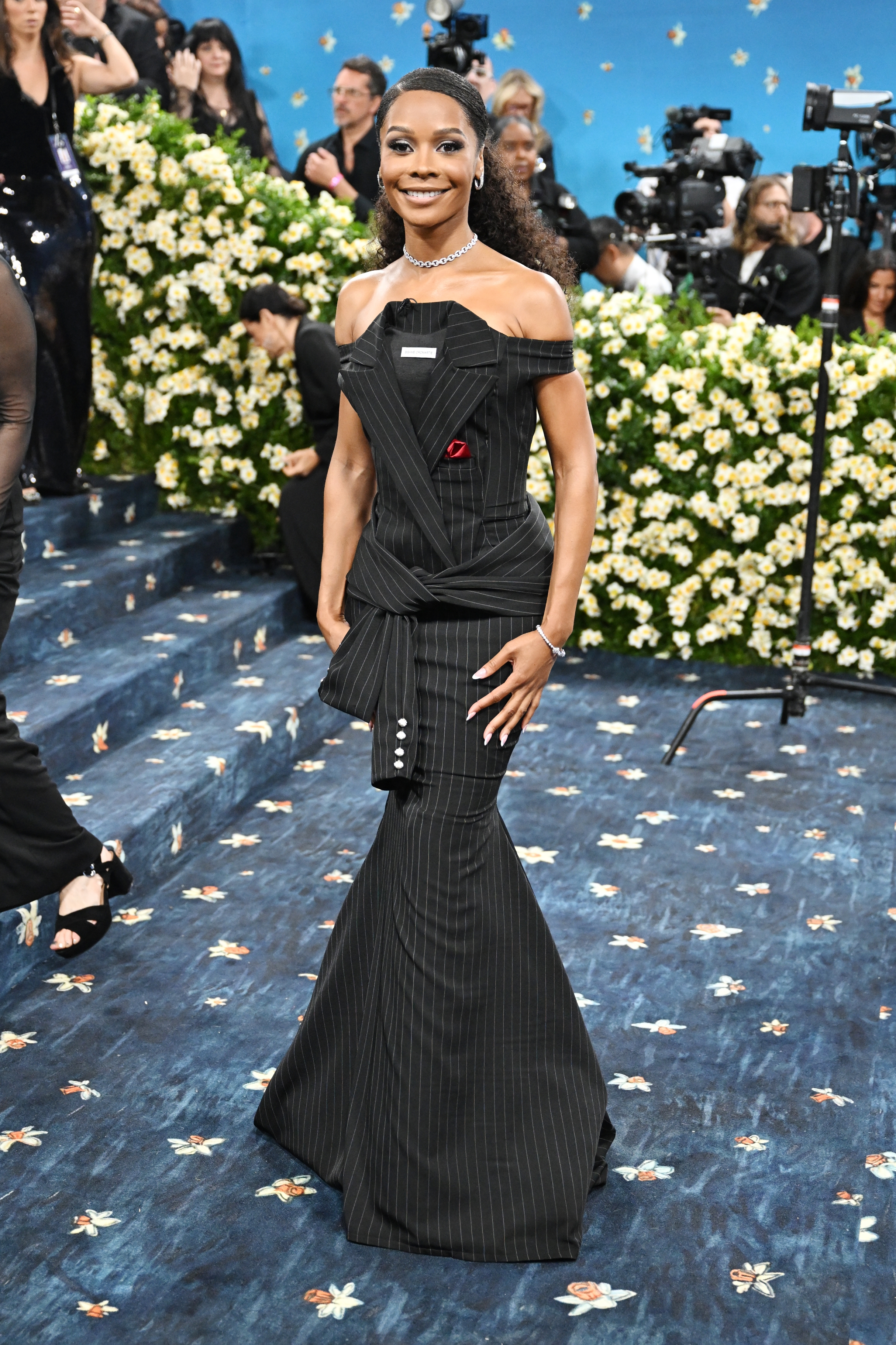 Person in an off-the-shoulder pinstripe gown on a floral-patterned red carpet. Background includes photographers and decorative flowers