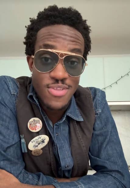 Person wearing sunglasses and a denim shirt with badges, looking at the camera with a neutral expression