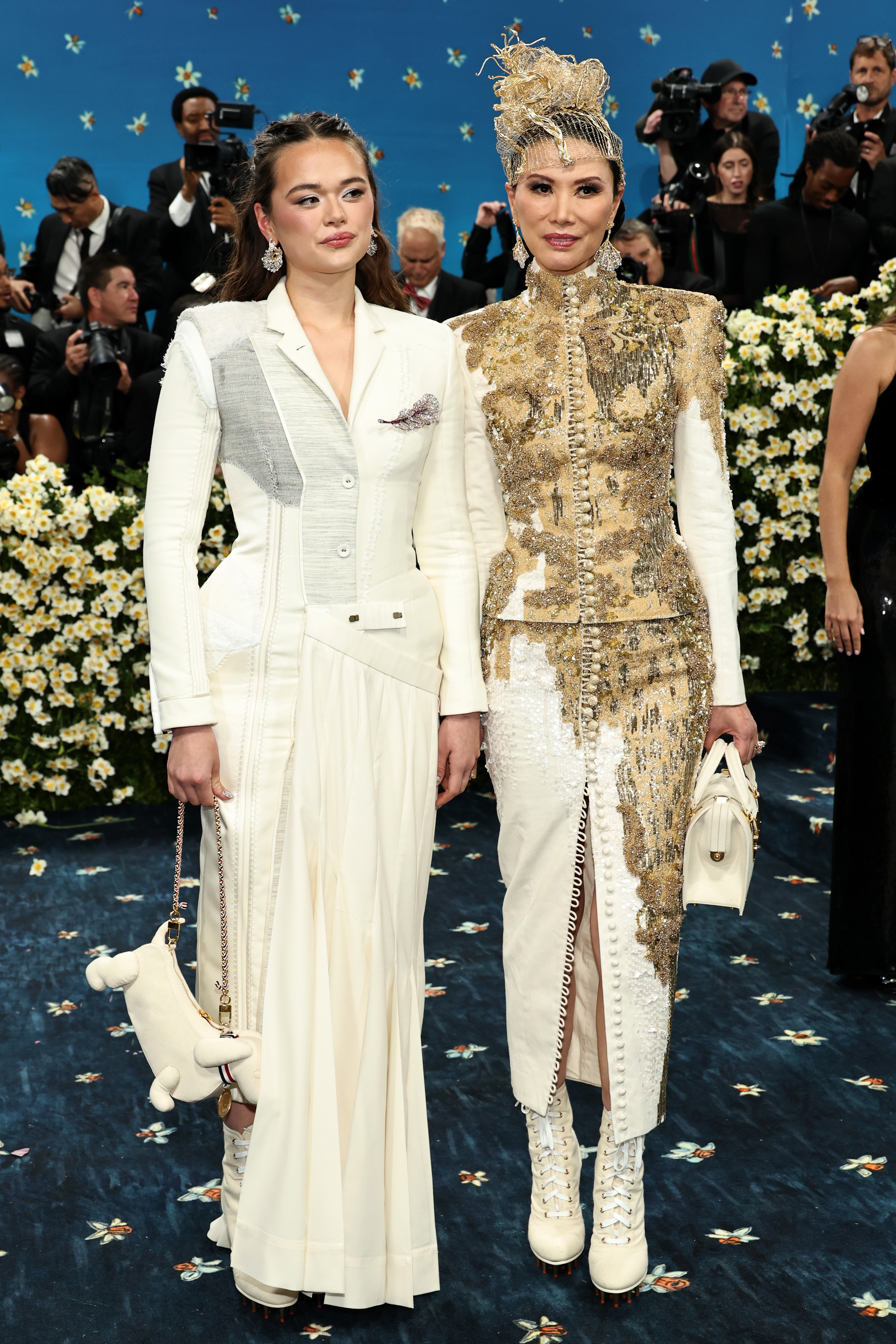 Two people on a red carpet. One wears a tailored white dress with layered elements, the other in an ornate gold and white ensemble with boots