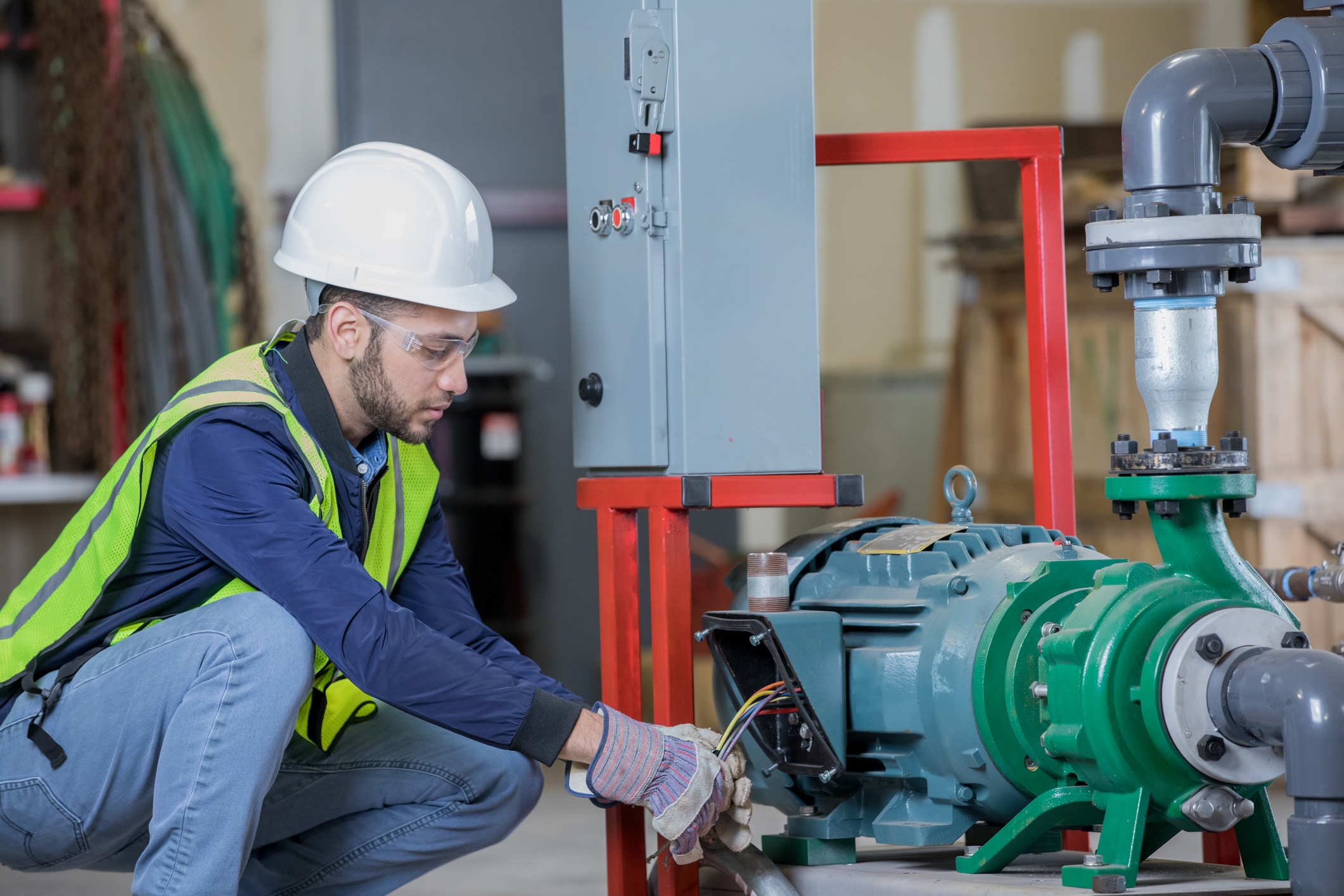 Person in safety gear and helmet works on industrial machinery, adjusting wires. Safety vest and gloves are visible. Industrial setup in the background