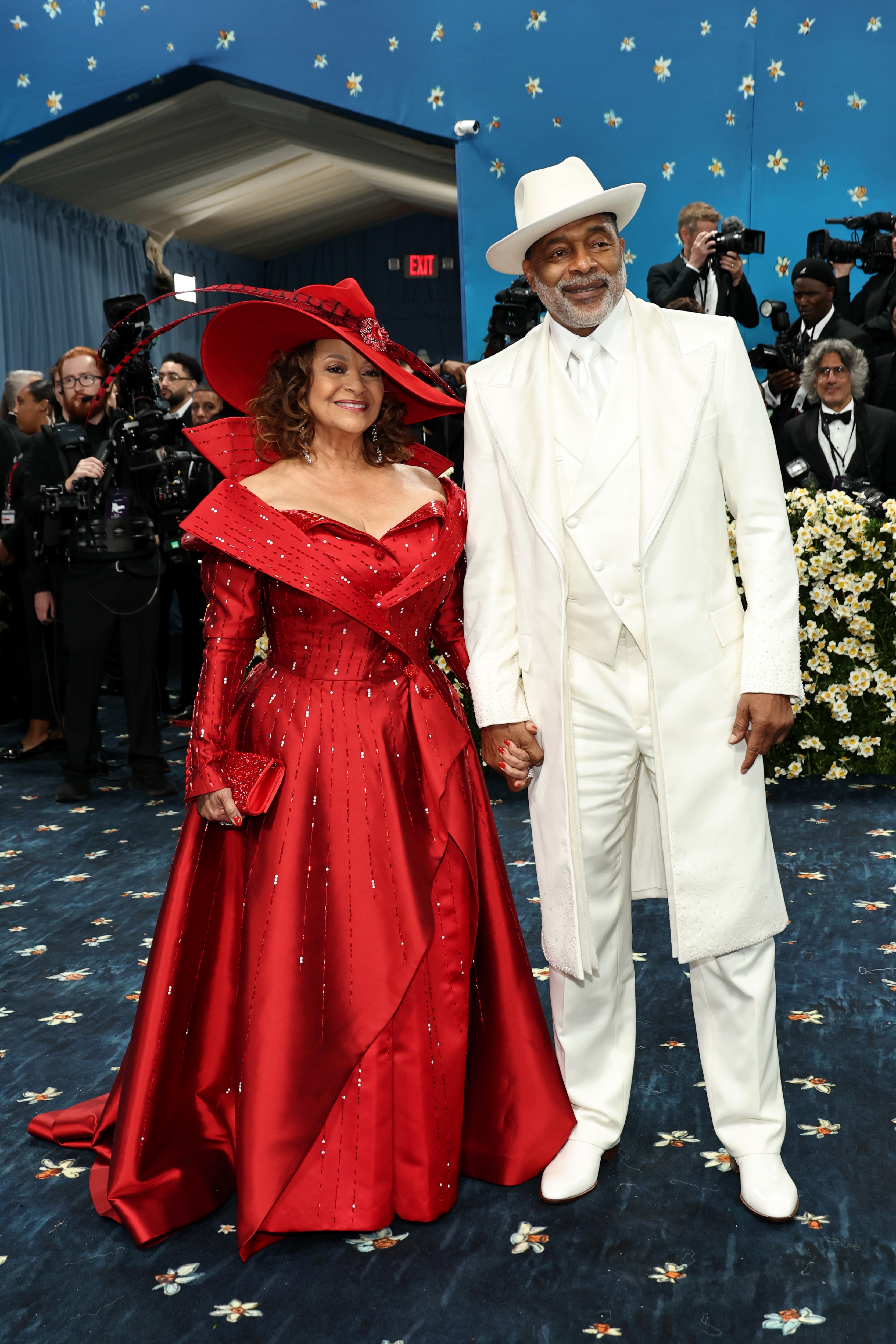 A couple posing on a star-patterned carpet; one in an ornate red gown and wide-brimmed hat, the other in a stylish white suit and hat