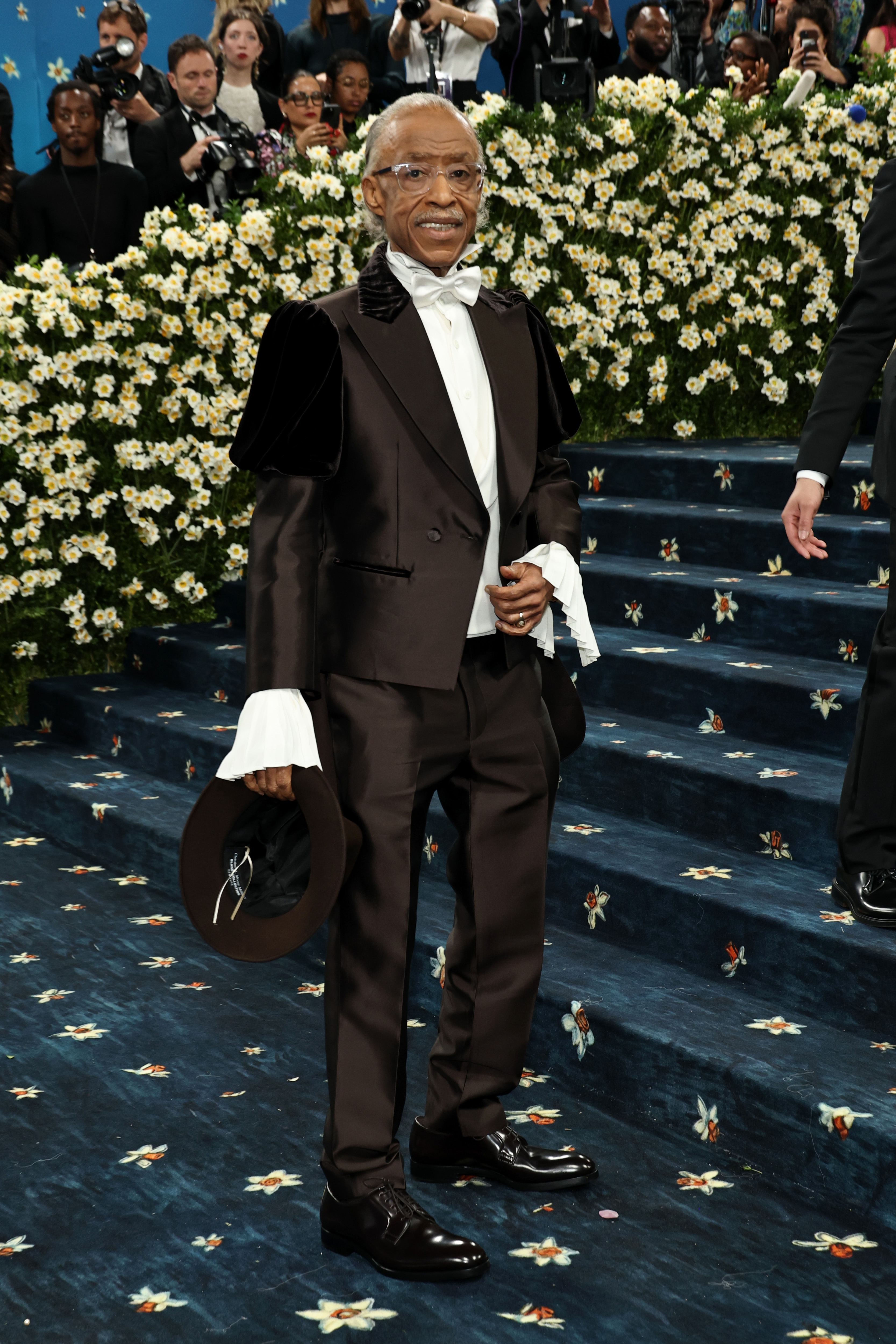 Man on red carpet in a tuxedo with ruffled shirt and bow tie, holding a hat, surrounded by flowers and photographers