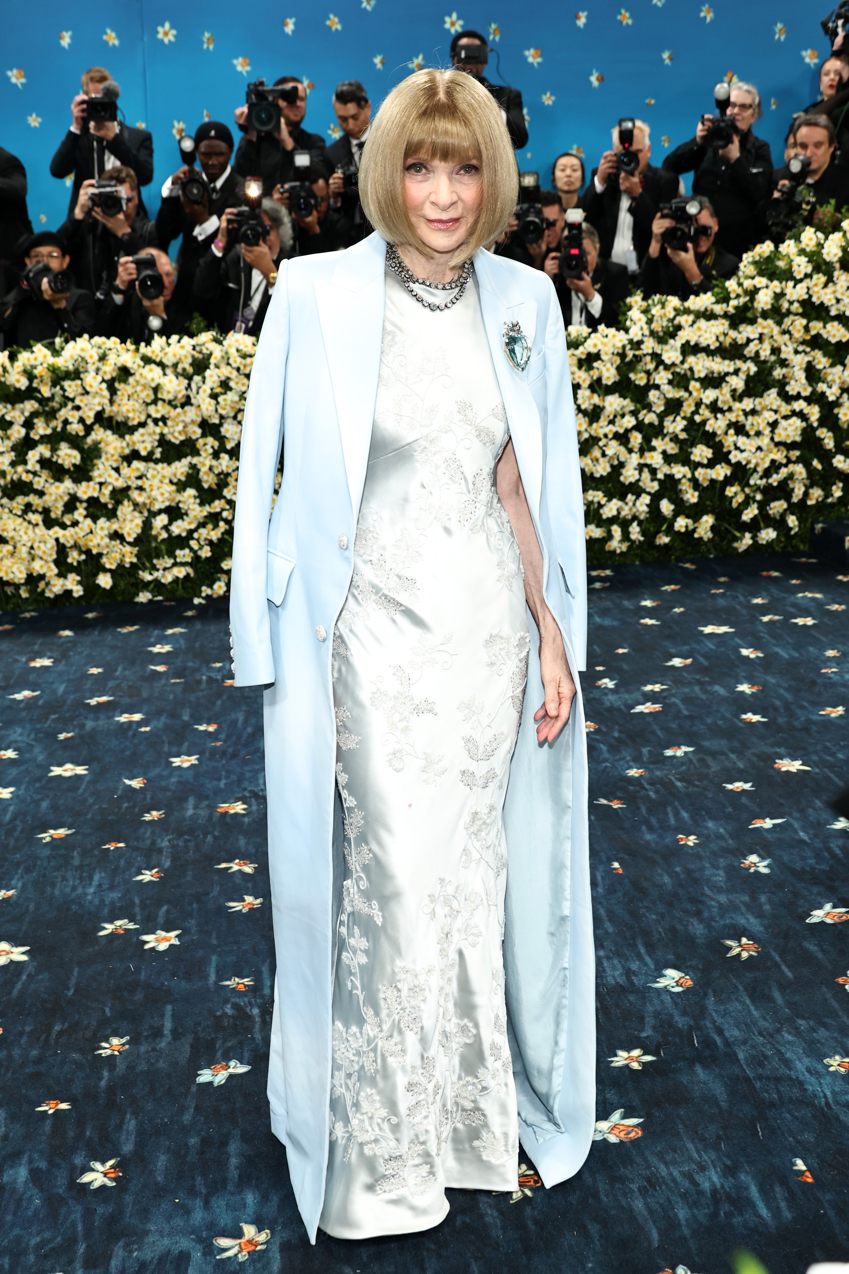 Person in an elegant embroidered gown with matching overcoat, surrounded by photographers on a formal event carpet