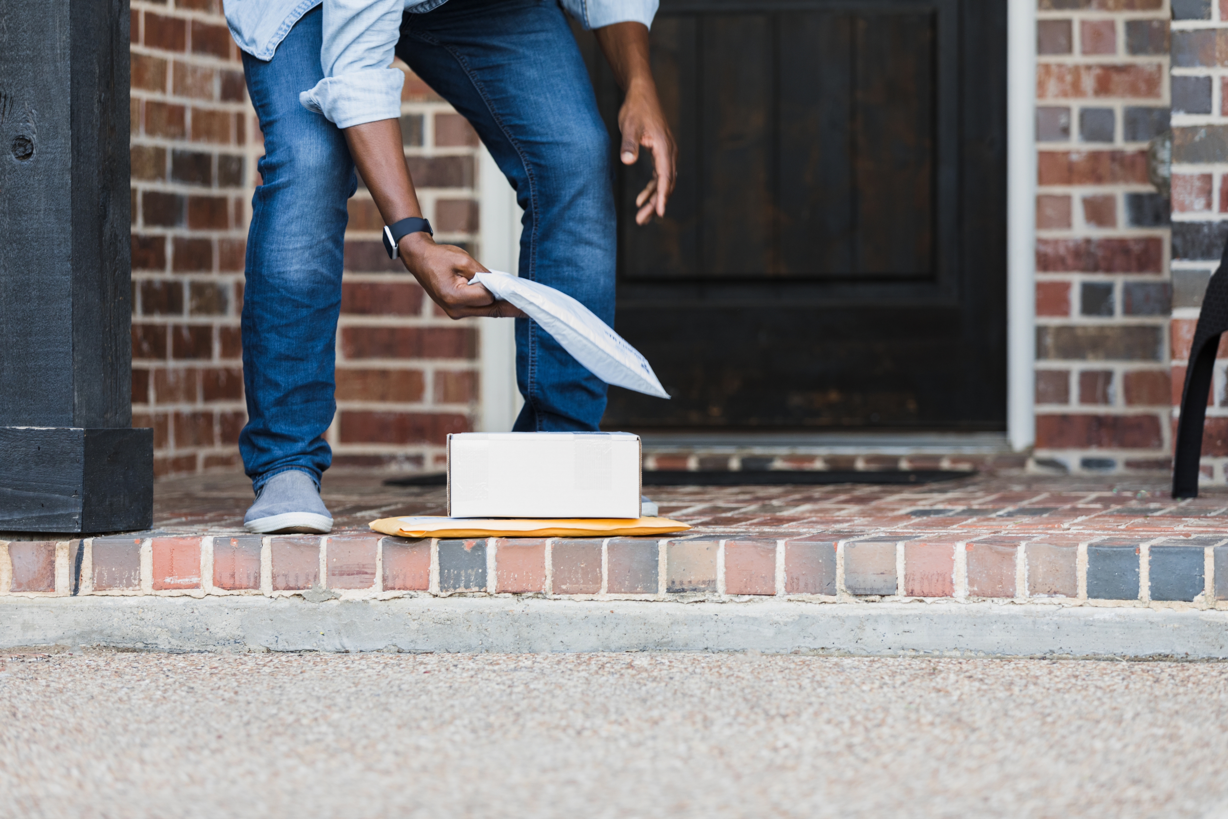 Person picking up mail from a doorstep, holding an envelope while bending over. Wearing casual jeans and gray shoes