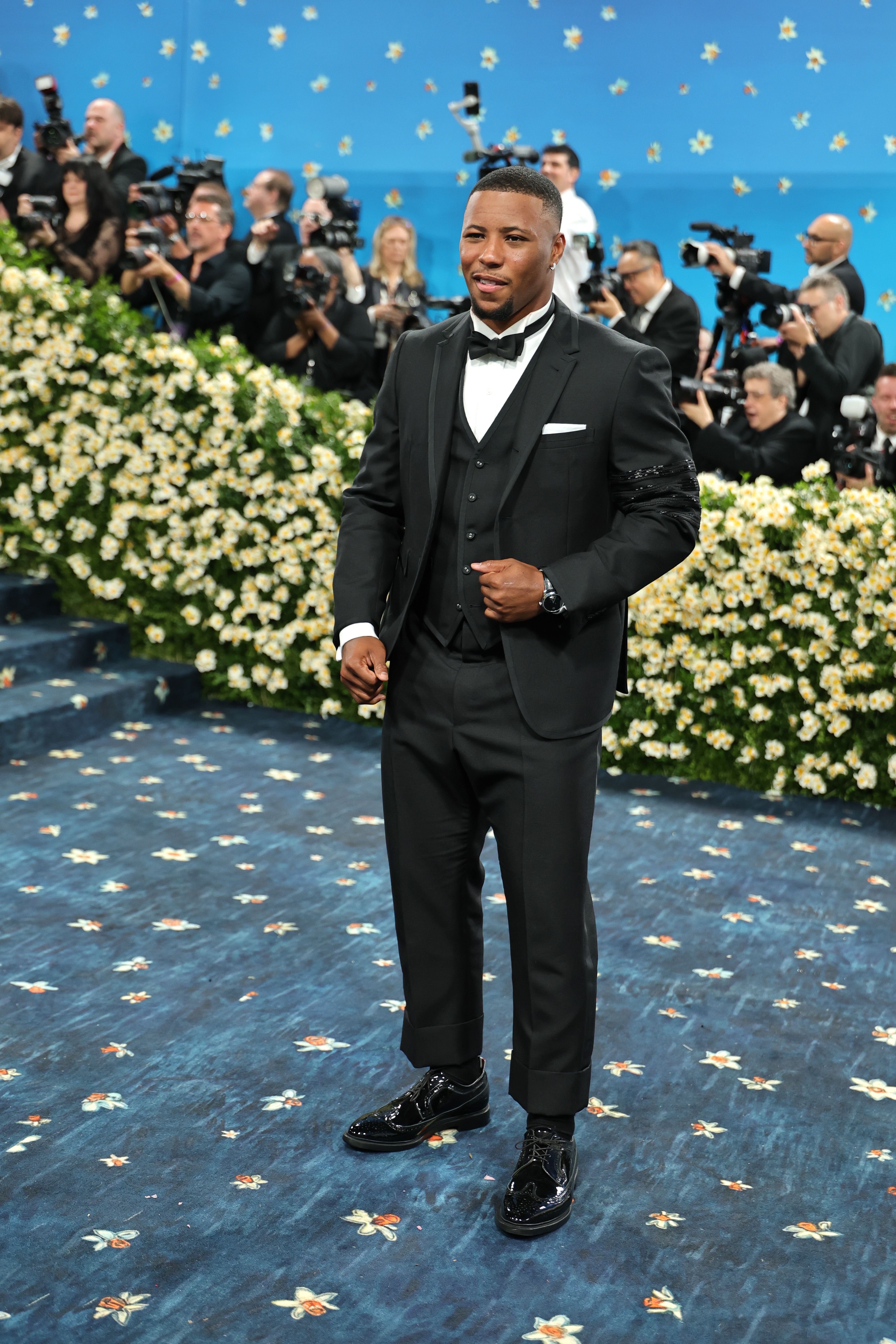 Person in a sharp suit with patent shoes poses on a floral-themed carpet at a formal event while photographers capture the moment