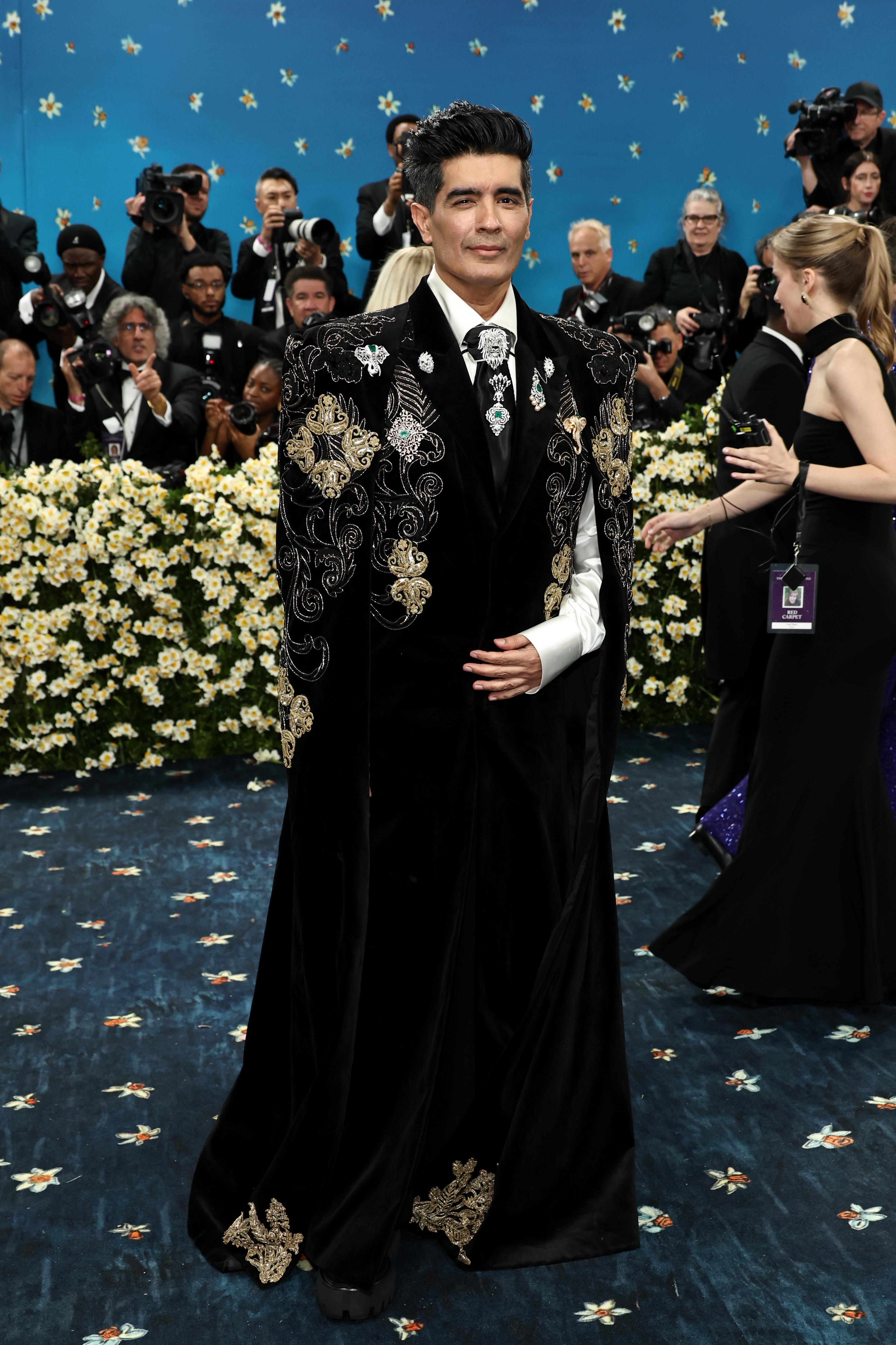 Person in embroidered cape and suit at a celebrity event, surrounded by photographers and attendees