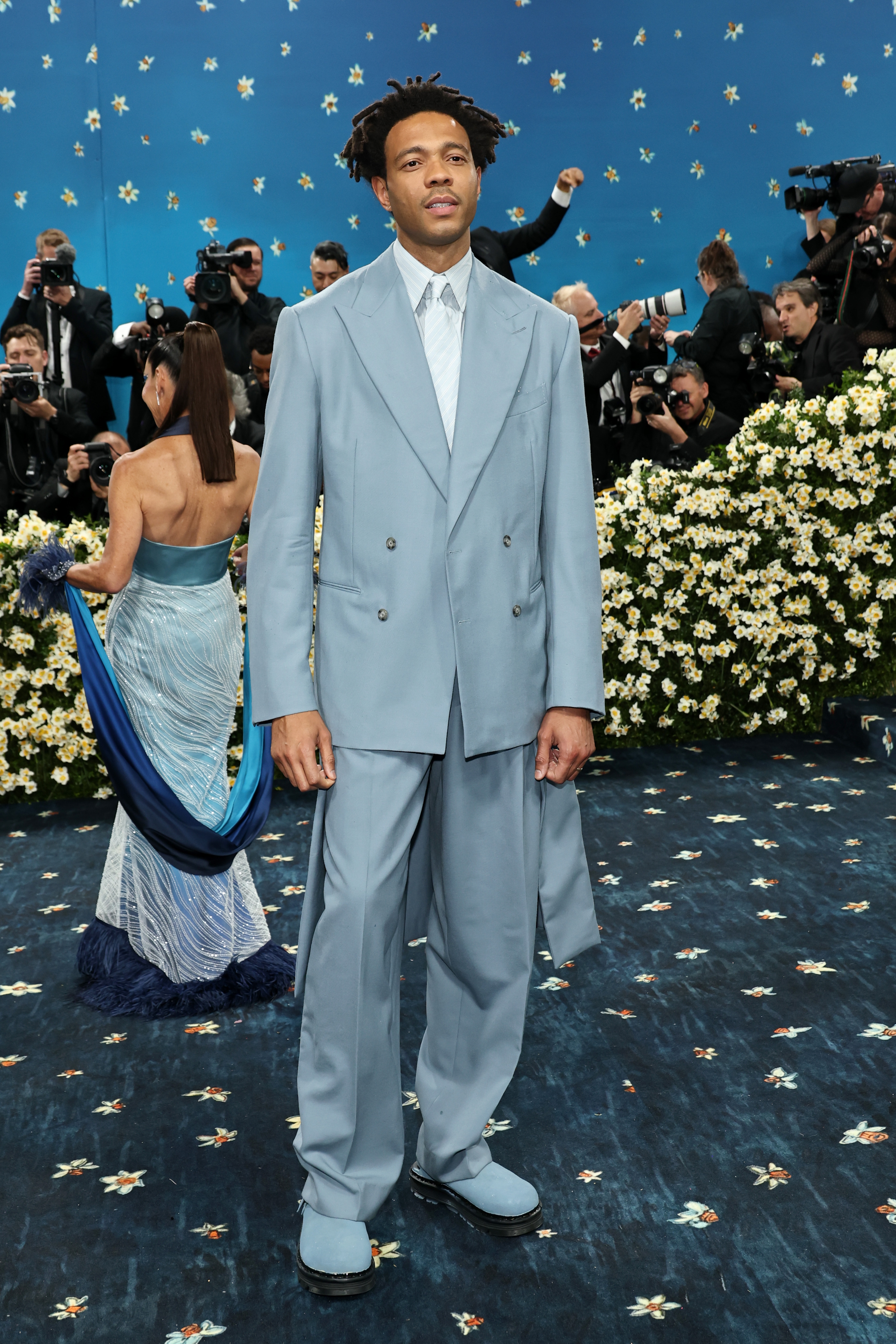 Person in an oversized suit on a floral-themed red carpet. Paparazzi and guests in elegant attire are in the background