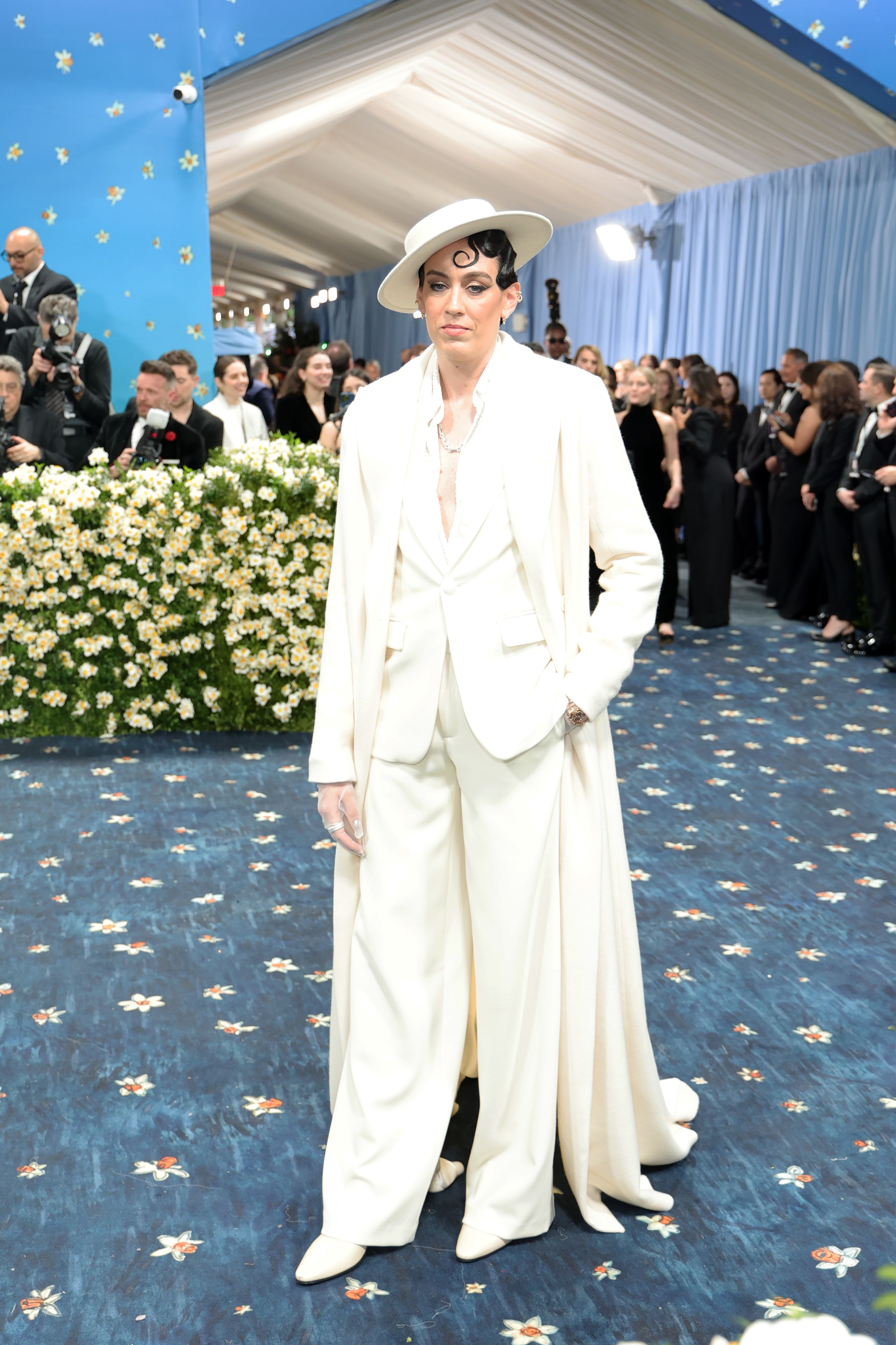 Person wearing a stylish white suit with a matching hat on a formal event's blue patterned carpet, surrounded by attendees