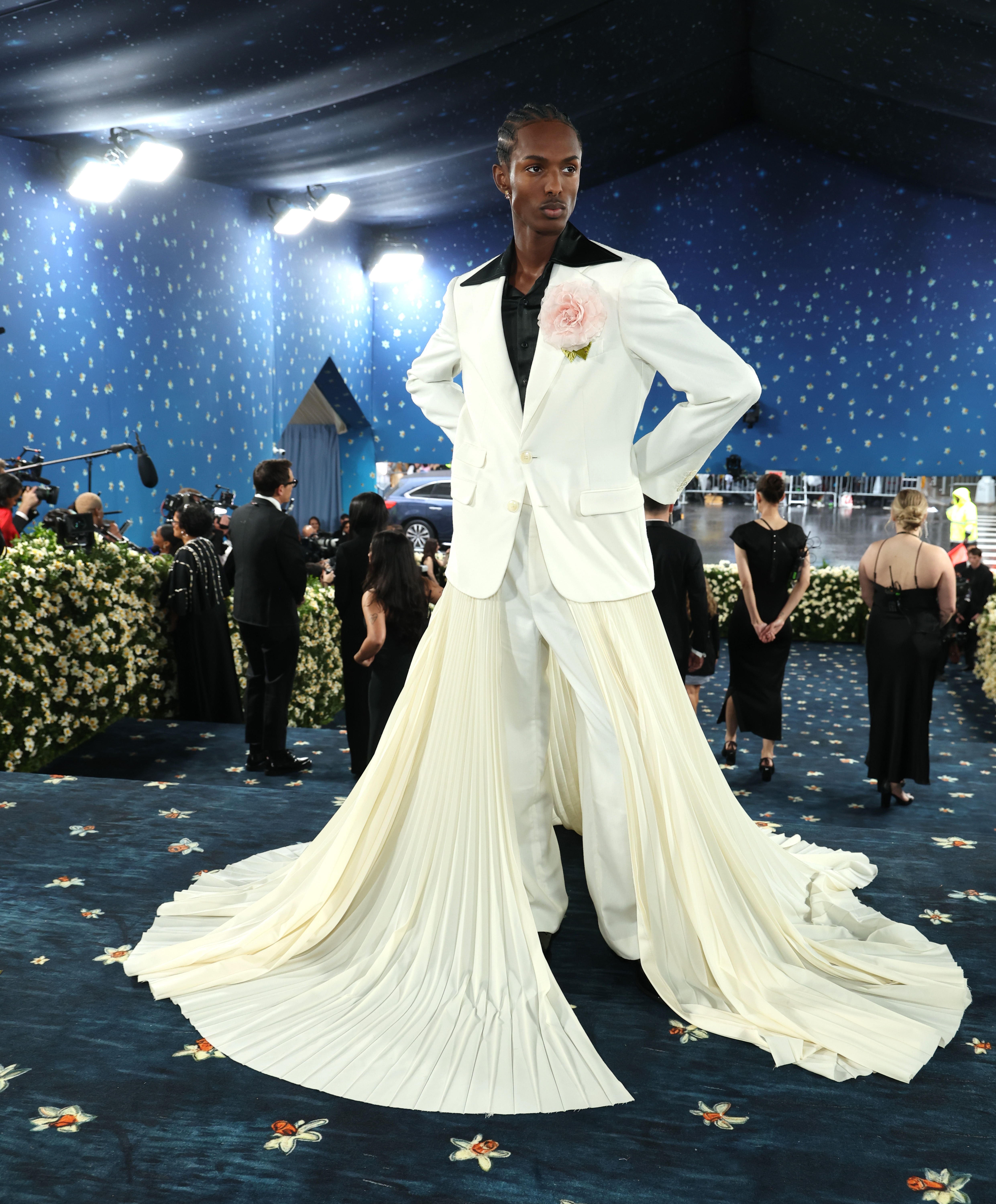 Person wearing an elegant white suit with a flowing pleated skirt on a starry-themed red carpet, surrounded by attendees and floral decorations