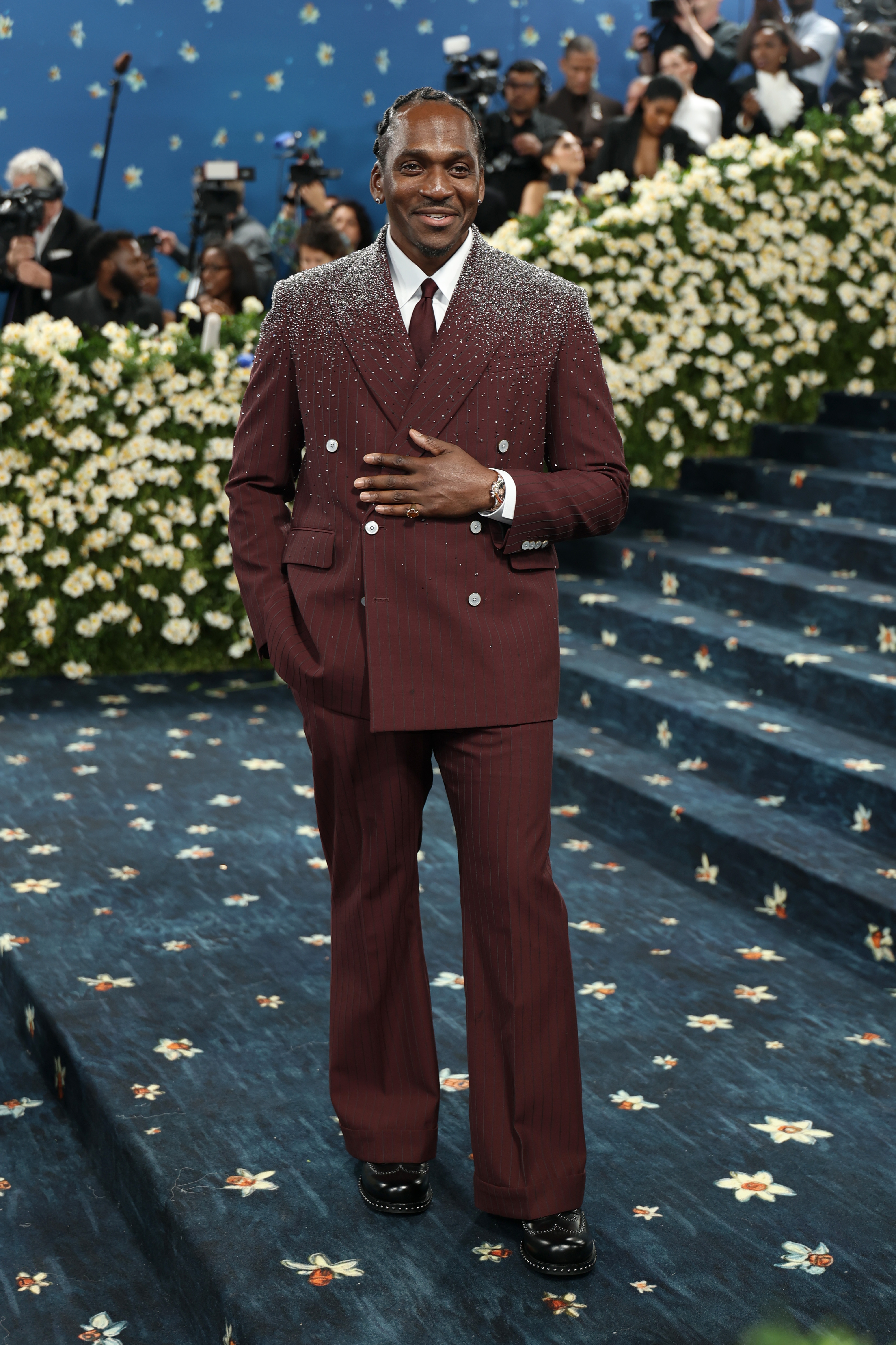 Person in a stylish suit with beaded embellishments stands on a floral-covered carpet, surrounded by photographers and onlookers