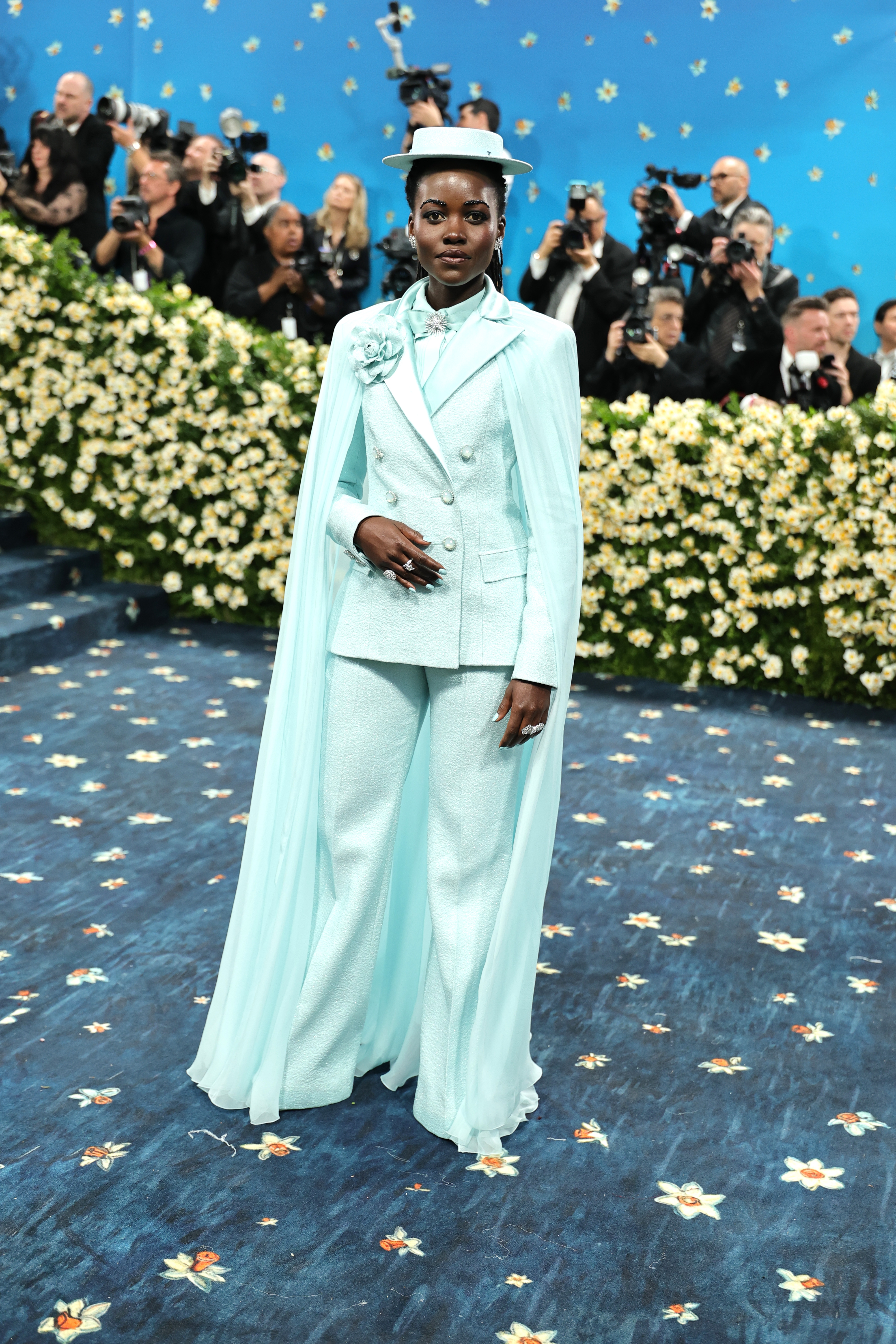Celebrity posing on floral-themed carpet in vintage-inspired suit with cape and hat, surrounded by photographers and floral backdrop