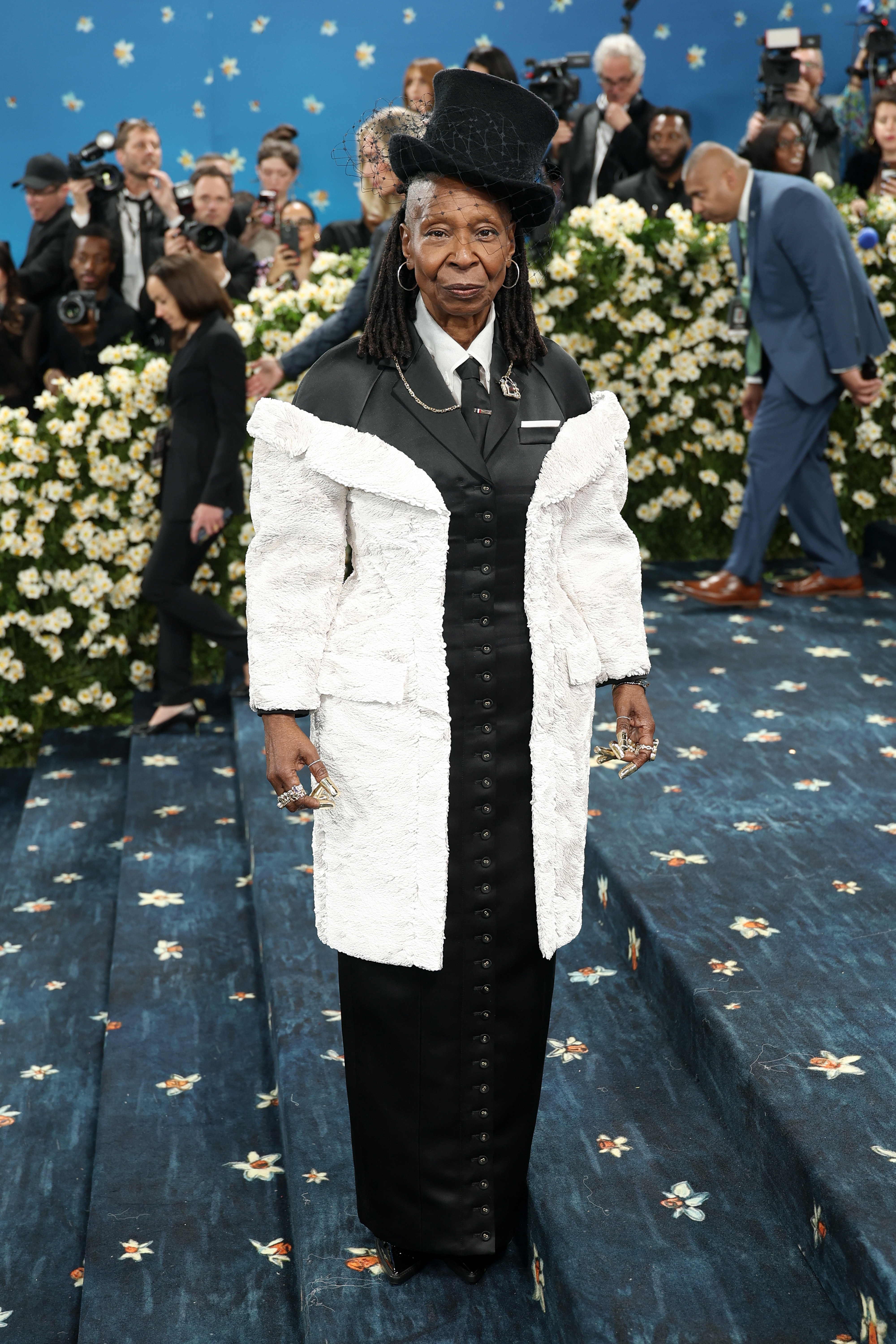 Person in a formal outfit with a long white coat and black hat, standing on an ornate staircase, surrounded by flowers