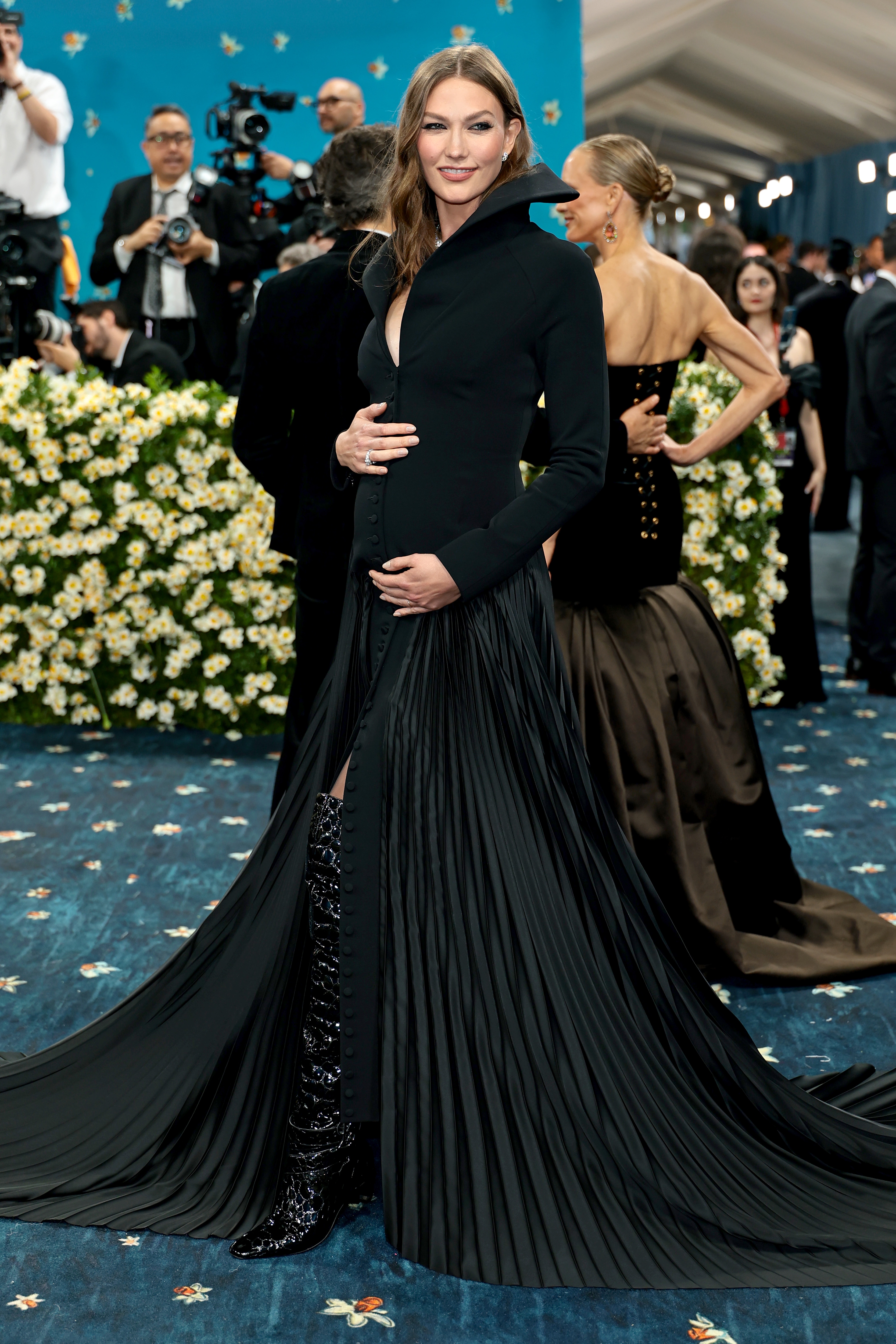 Person in elegant long coat dress with high collar and floor-length train, poses on a blue-patterned carpet at a formal event, surrounded by flowers
