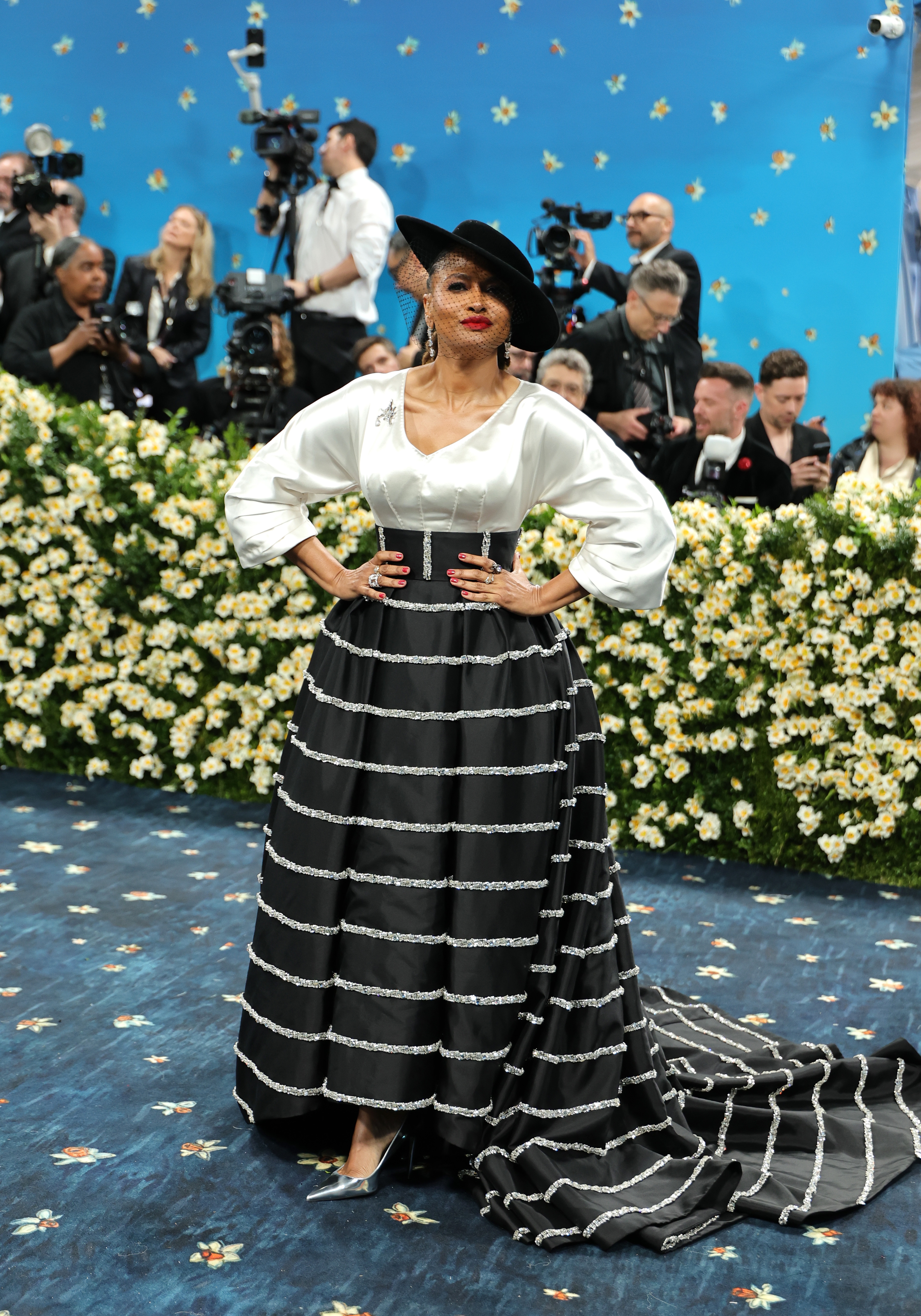 Celebrity on red carpet wearing a wide-brimmed hat and a striped gown with a full skirt, posing confidently surrounded by media and photographers