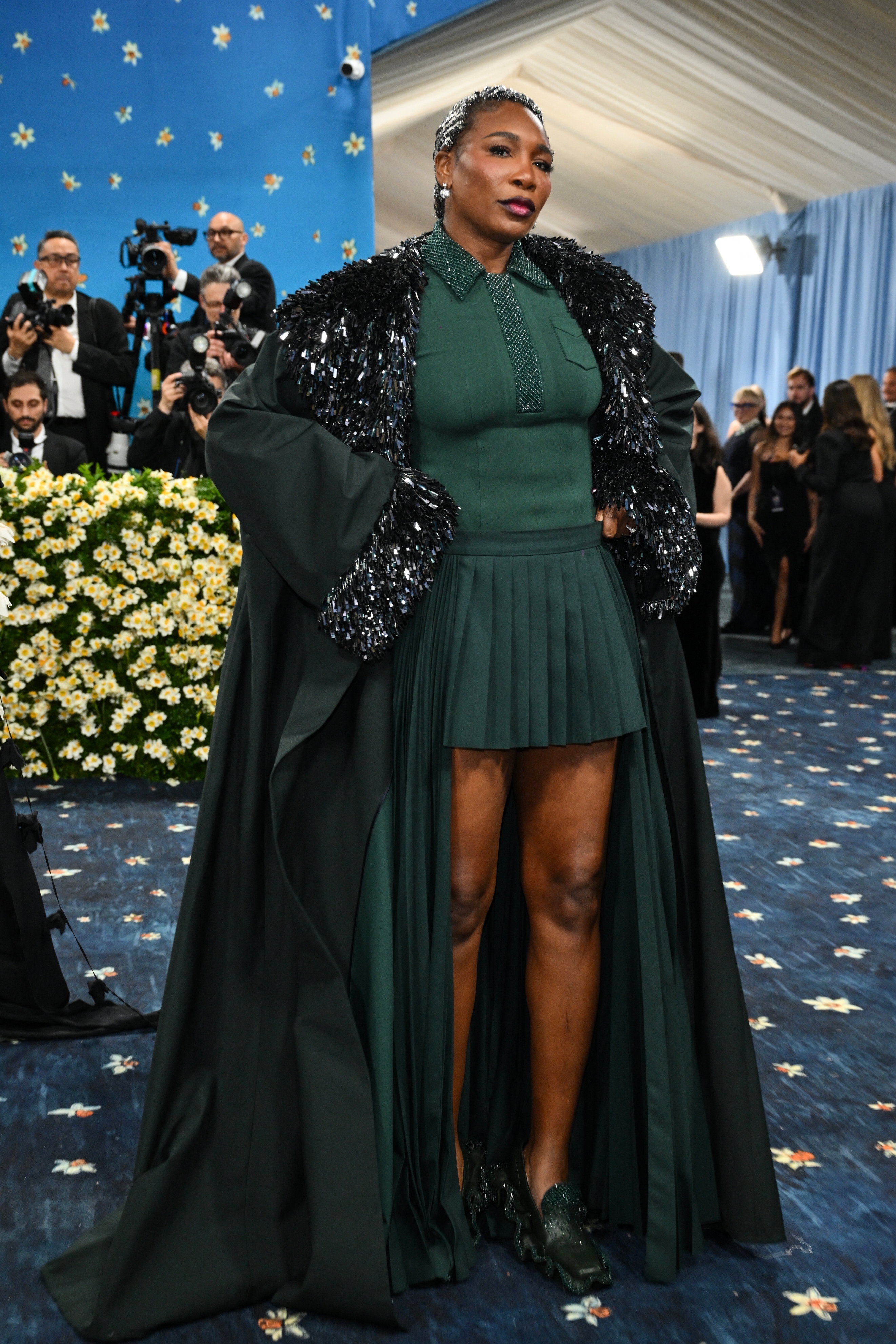 Person in a chic gown with sparkling details and a cape on a formal event carpet, surrounded by attendees and photographers