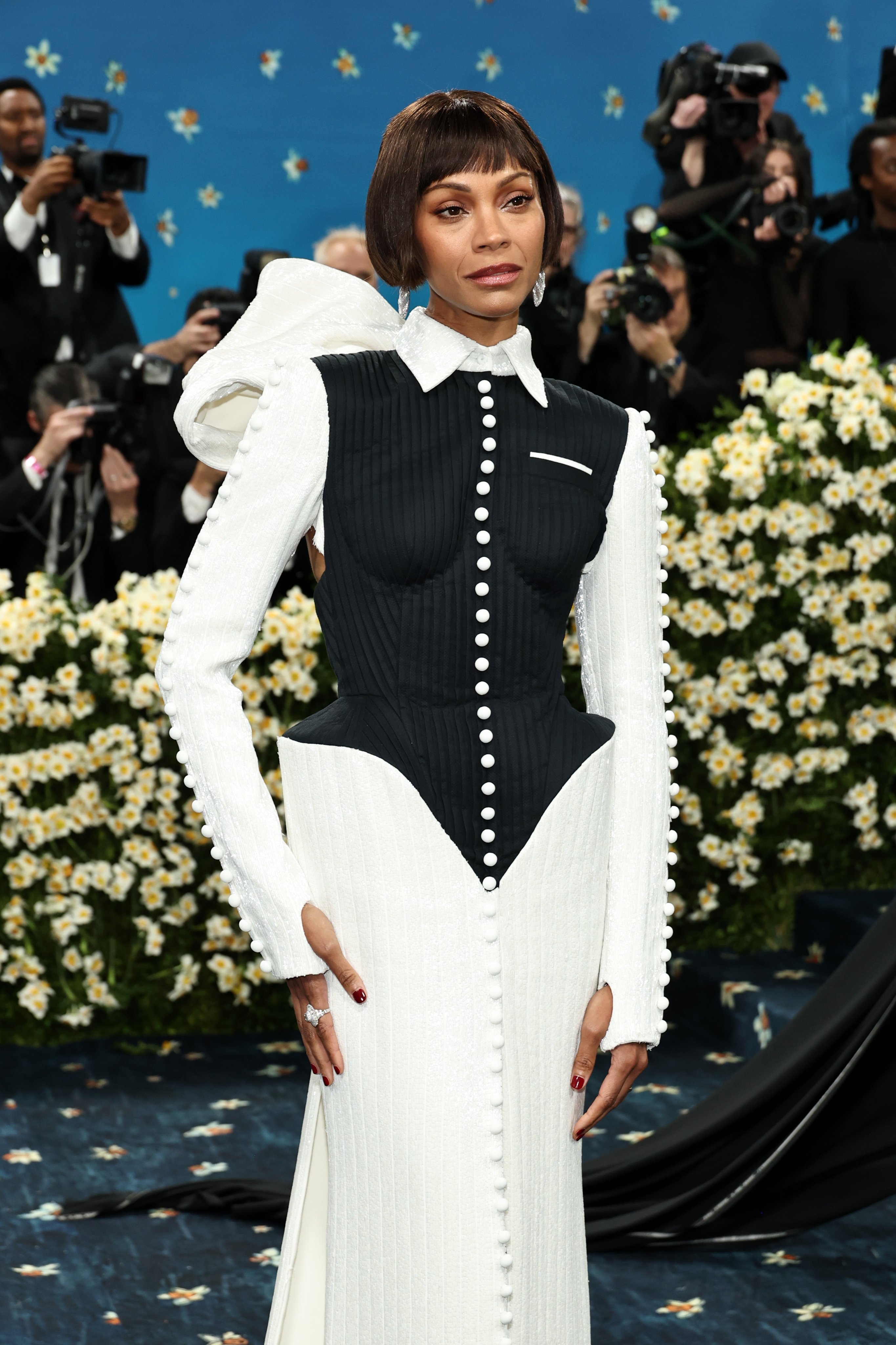 Persona no identificada con vestido de gala negro y blanco con botones y detalle estructural en un evento concurrido; cámaras al fondo