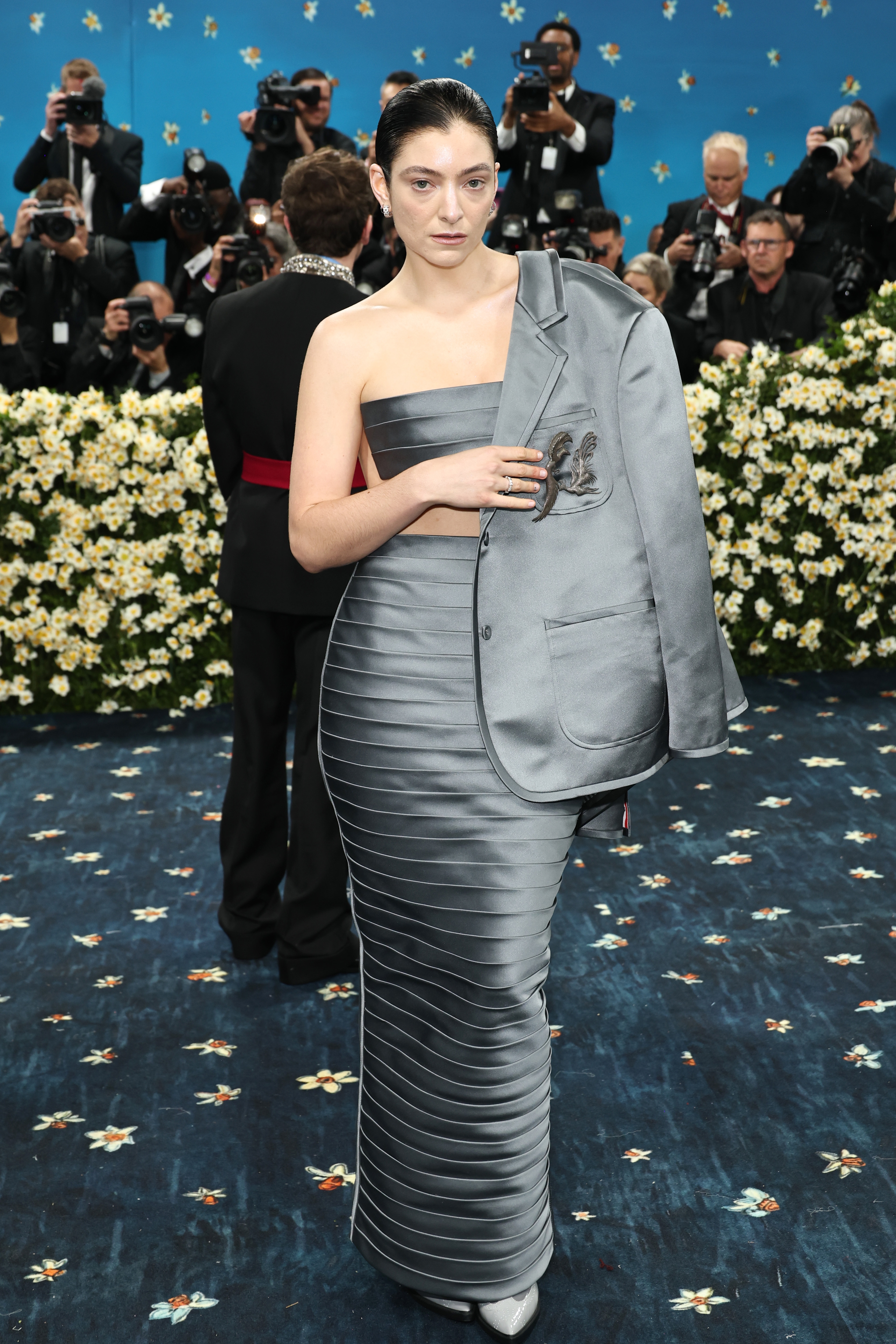Person in a strapless, ribbed gown with a jacket over one shoulder, posing on a floral-themed event carpet surrounded by photographers