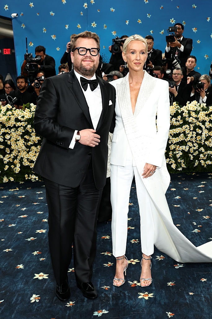 Two people in formal attire pose on a star-patterned carpet, surrounded by photographers. One wears a tuxedo, the other a white suit with a train