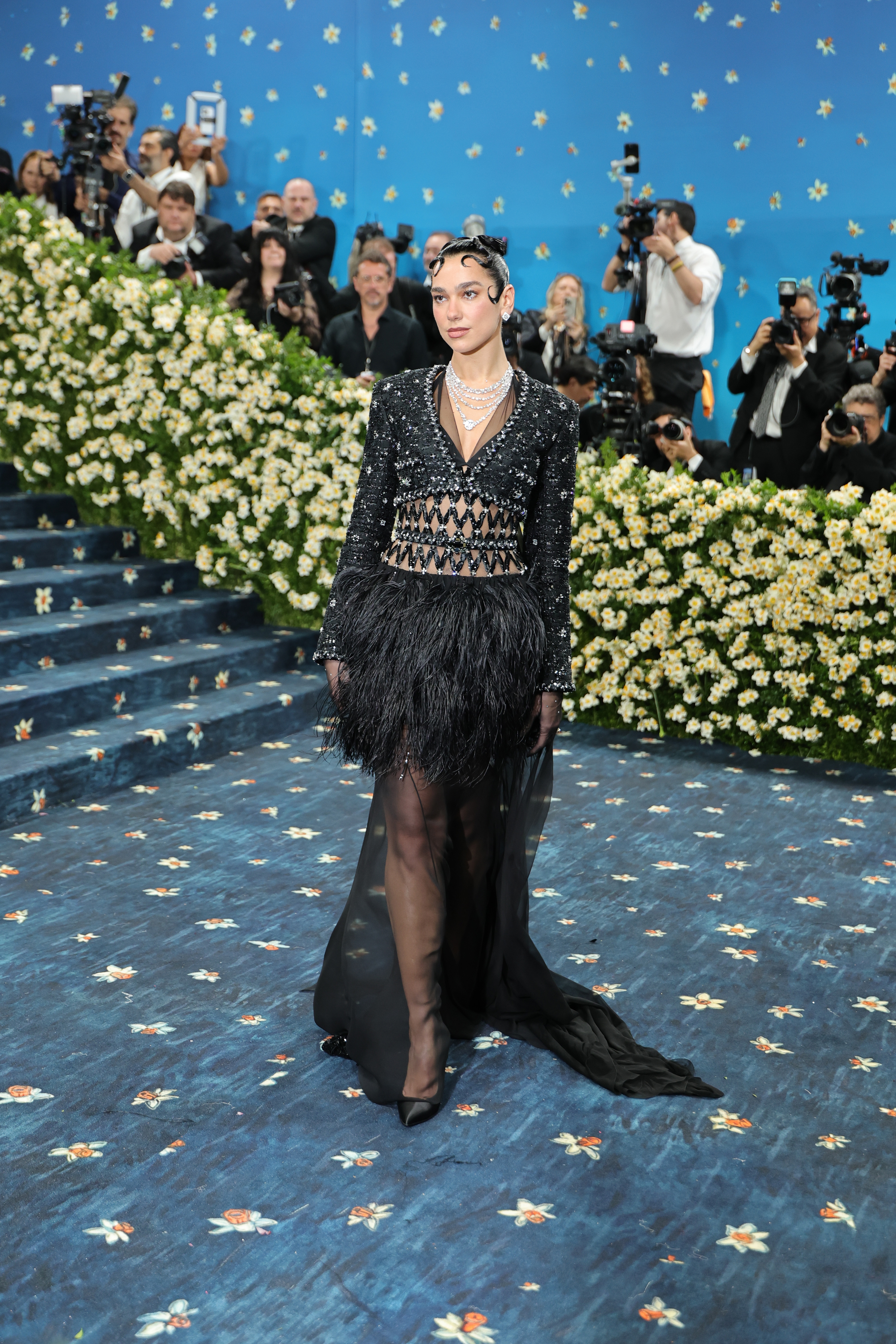 Person in a black, sparkling gown with a feathered skirt on a floral-decorated carpet, posing for photographers