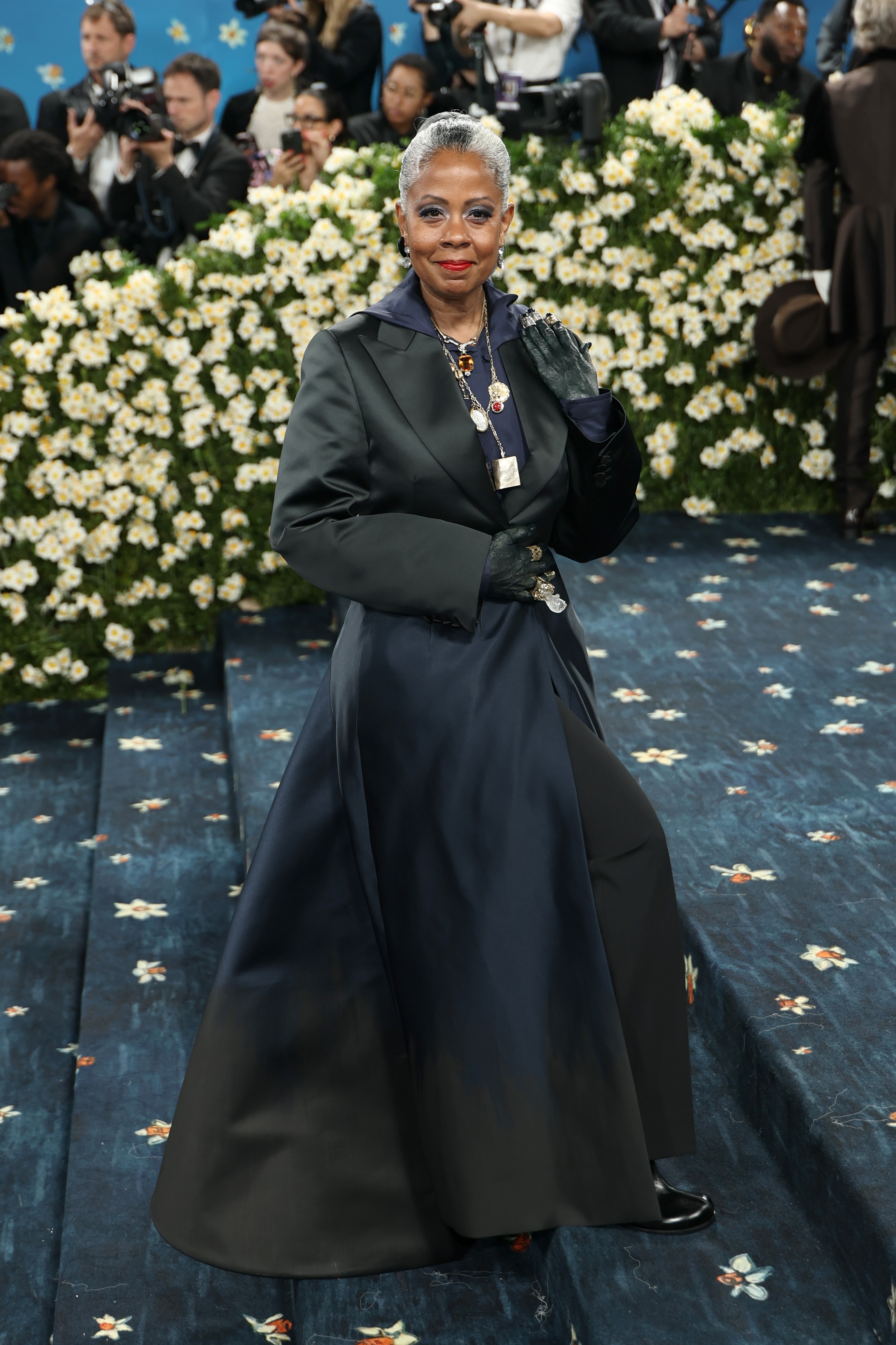 A person in an elegant long coat and gown poses on a flower-adorned staircase, surrounded by photographers