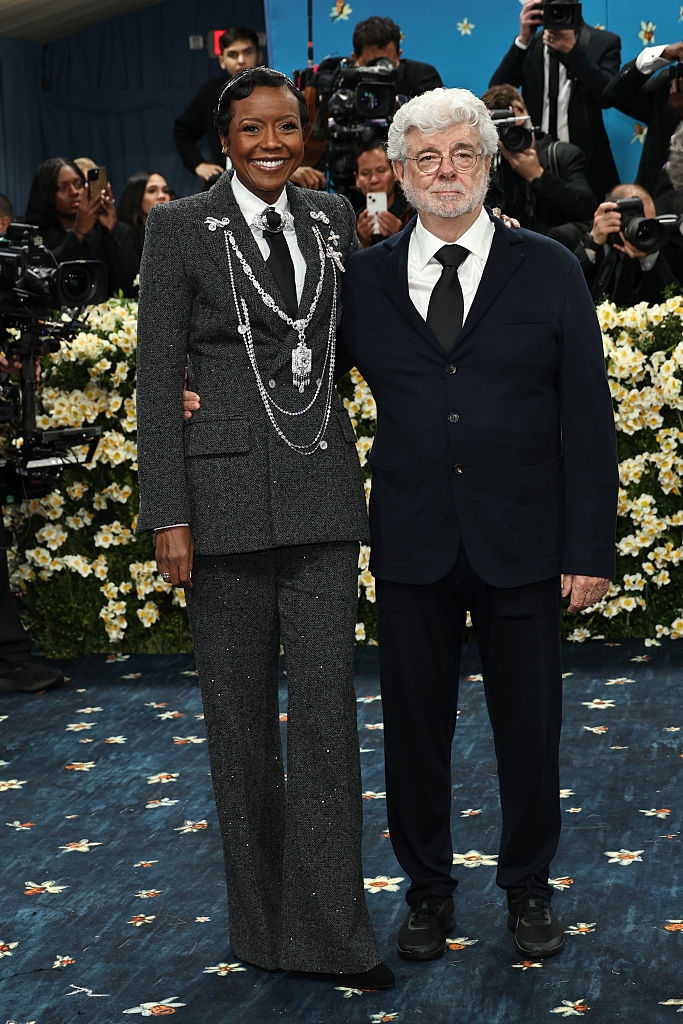 Mellody wears a textured suit with chain accessories, George wears a classic dark suit and tie