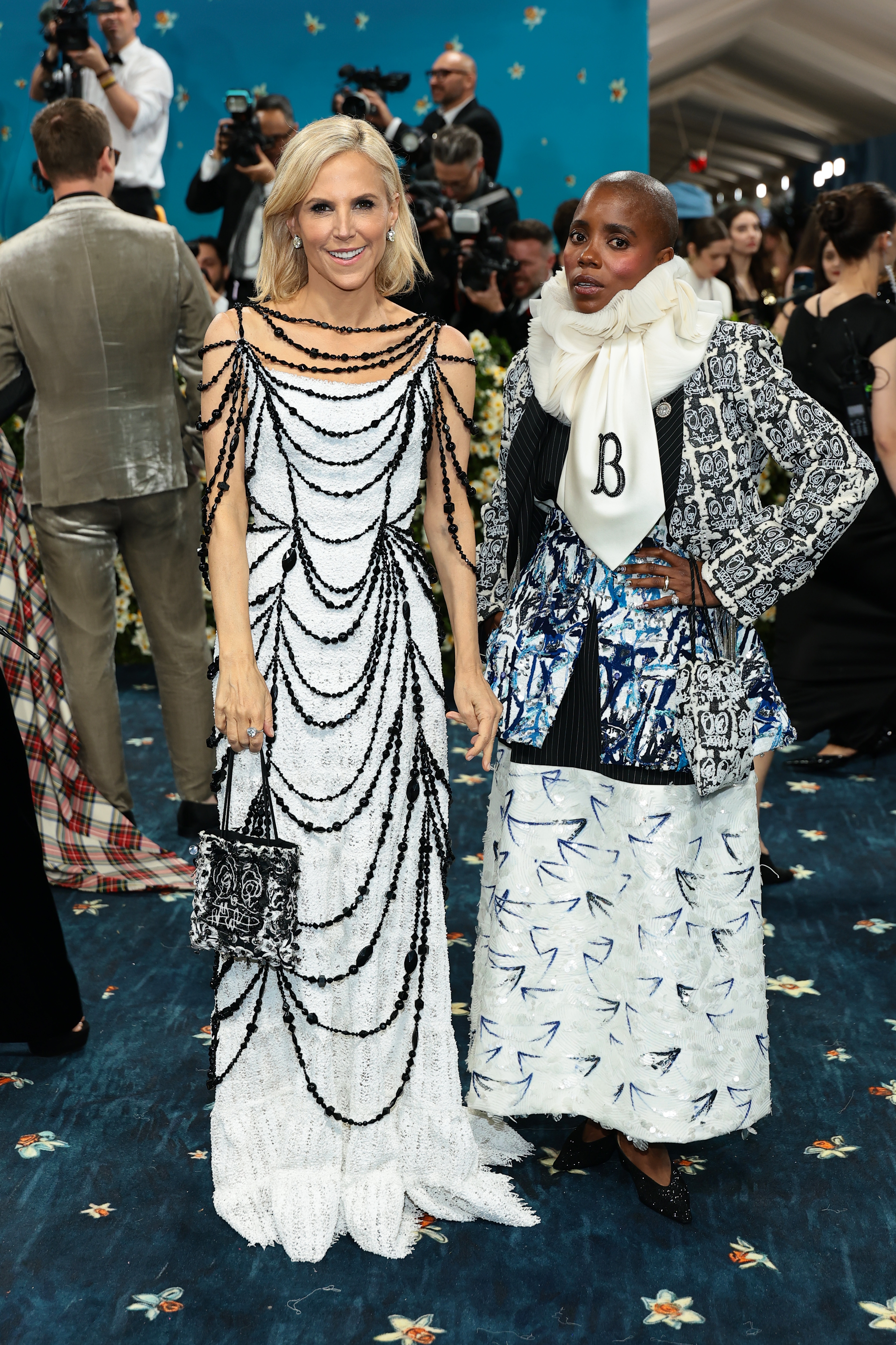 Two individuals on a red carpet: one in a beaded dress with draped chains, the other in a patterned blazer, scarf, and skirt, posing for photos