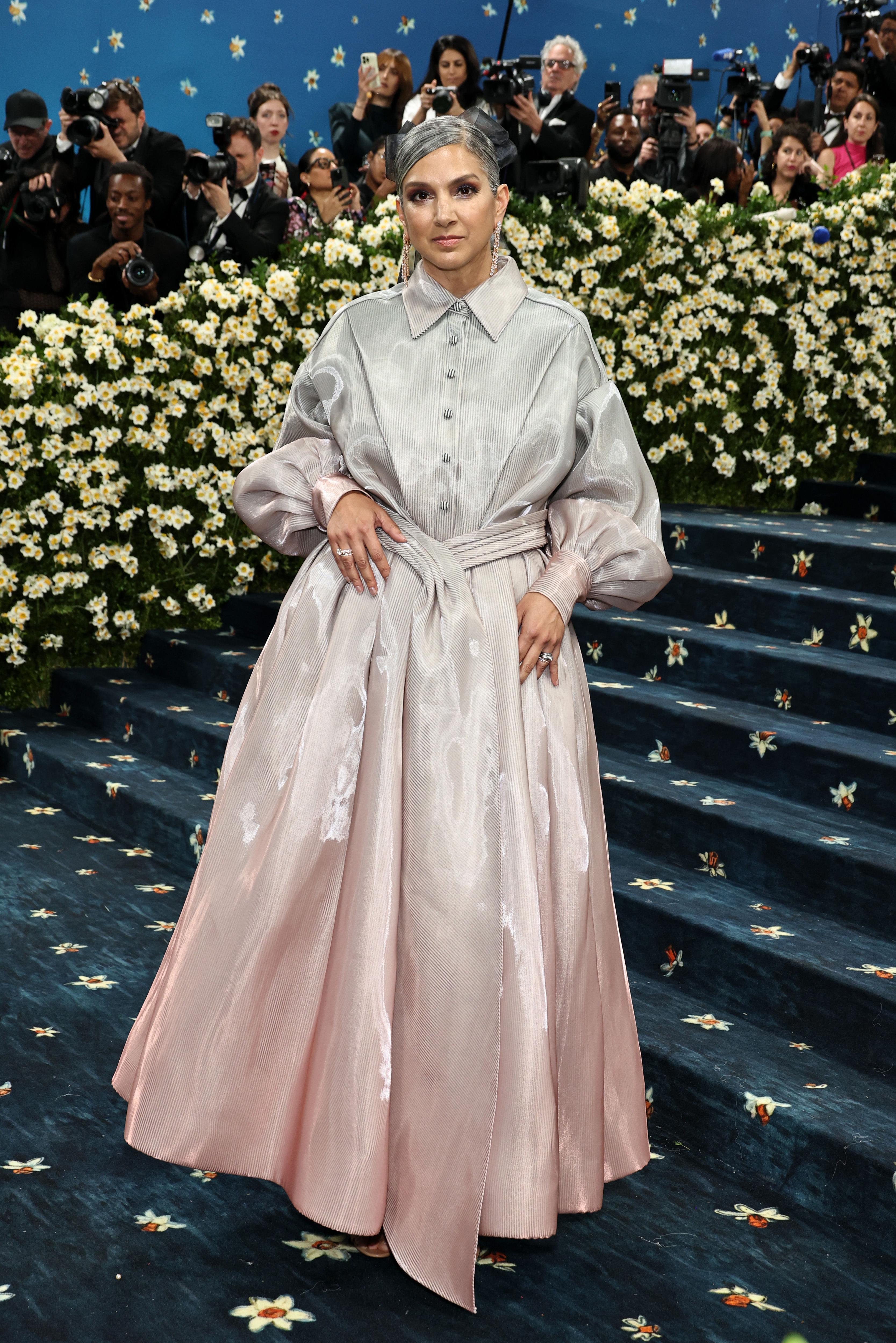 Person in a shiny, flowing gown with a cinched waist, posing on a floral-themed red carpet surrounded by photographers