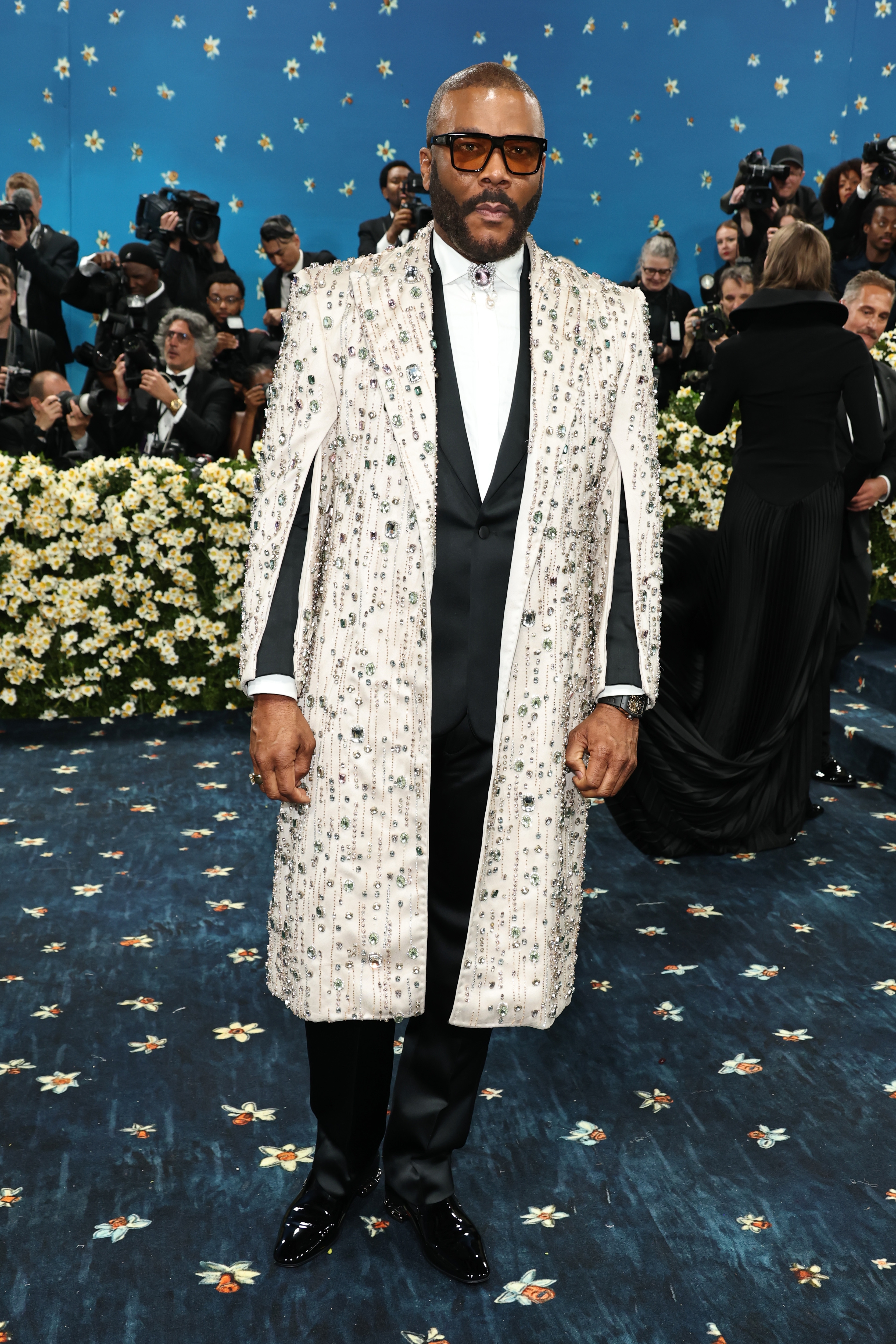 A man on the red carpet wears a patterned, knee-length coat over a formal black suit with a white shirt, surrounded by photographers