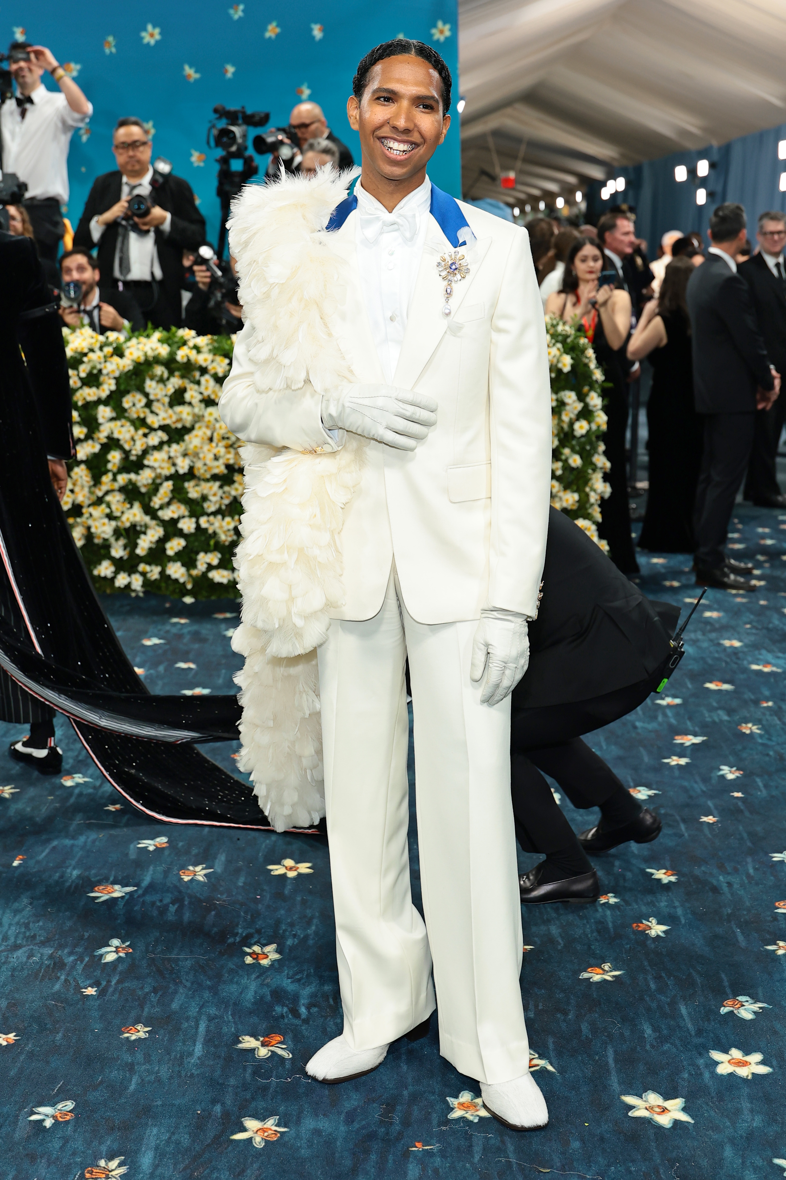 Person in a formal white suit with extravagant feathered cape, smiling on a glamorous event carpet. Crowd and photographers in the background