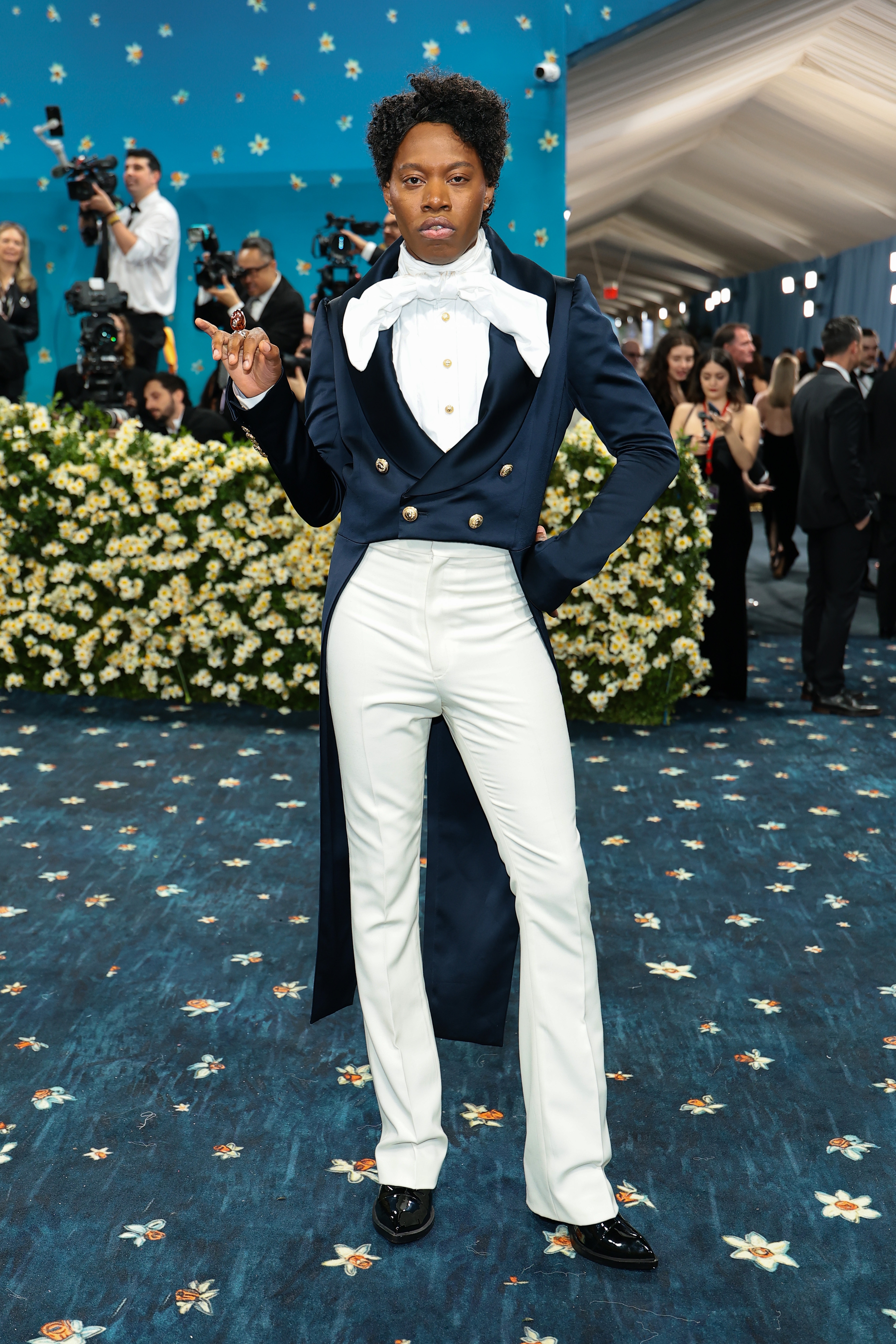 Person in a ruffled white blouse and navy coat with white pants, standing confidently on a floral carpet with photographers in the background