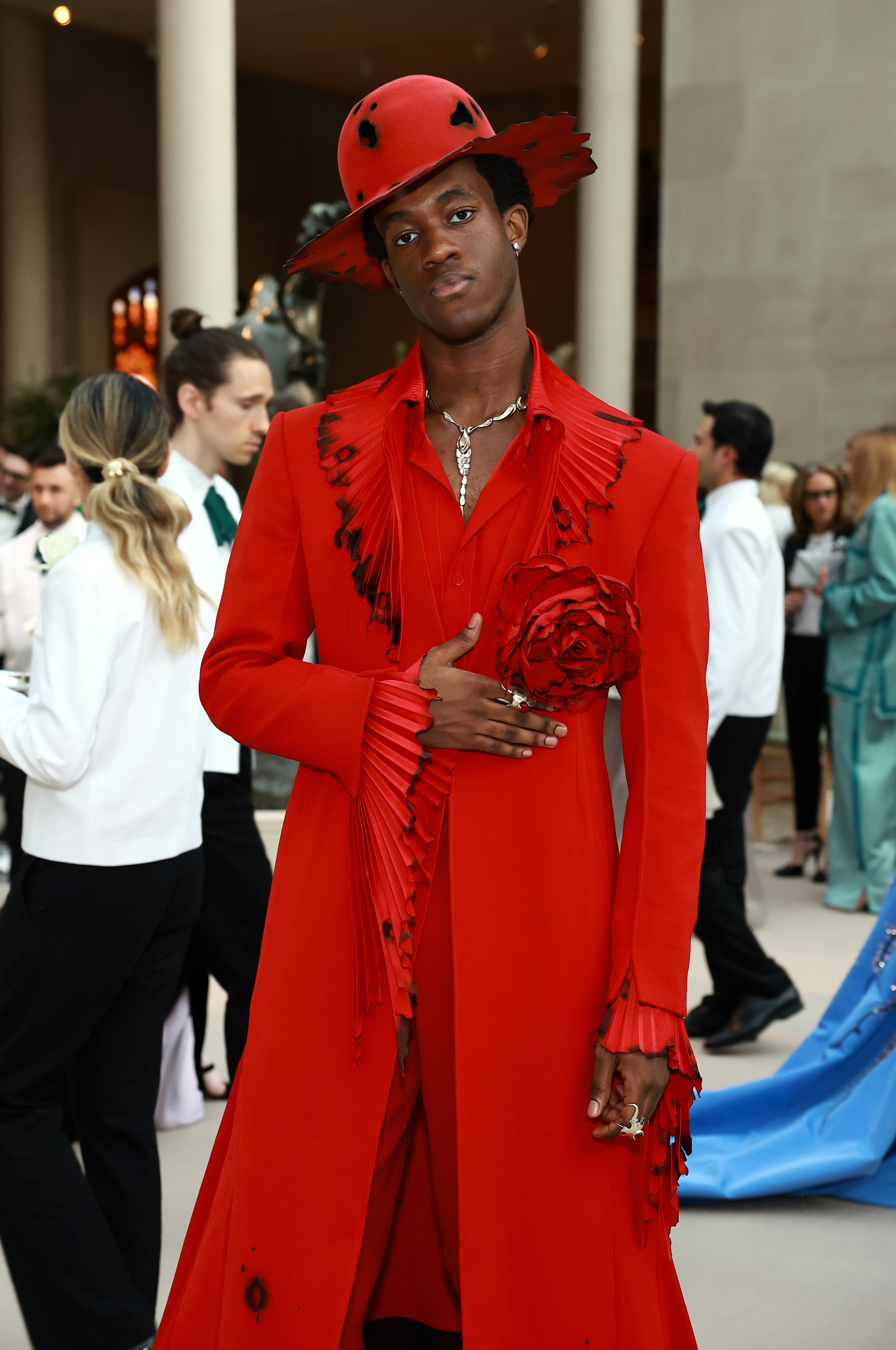 Person in an elaborate red outfit with a matching hat and large fabric rose detail, posing at an event