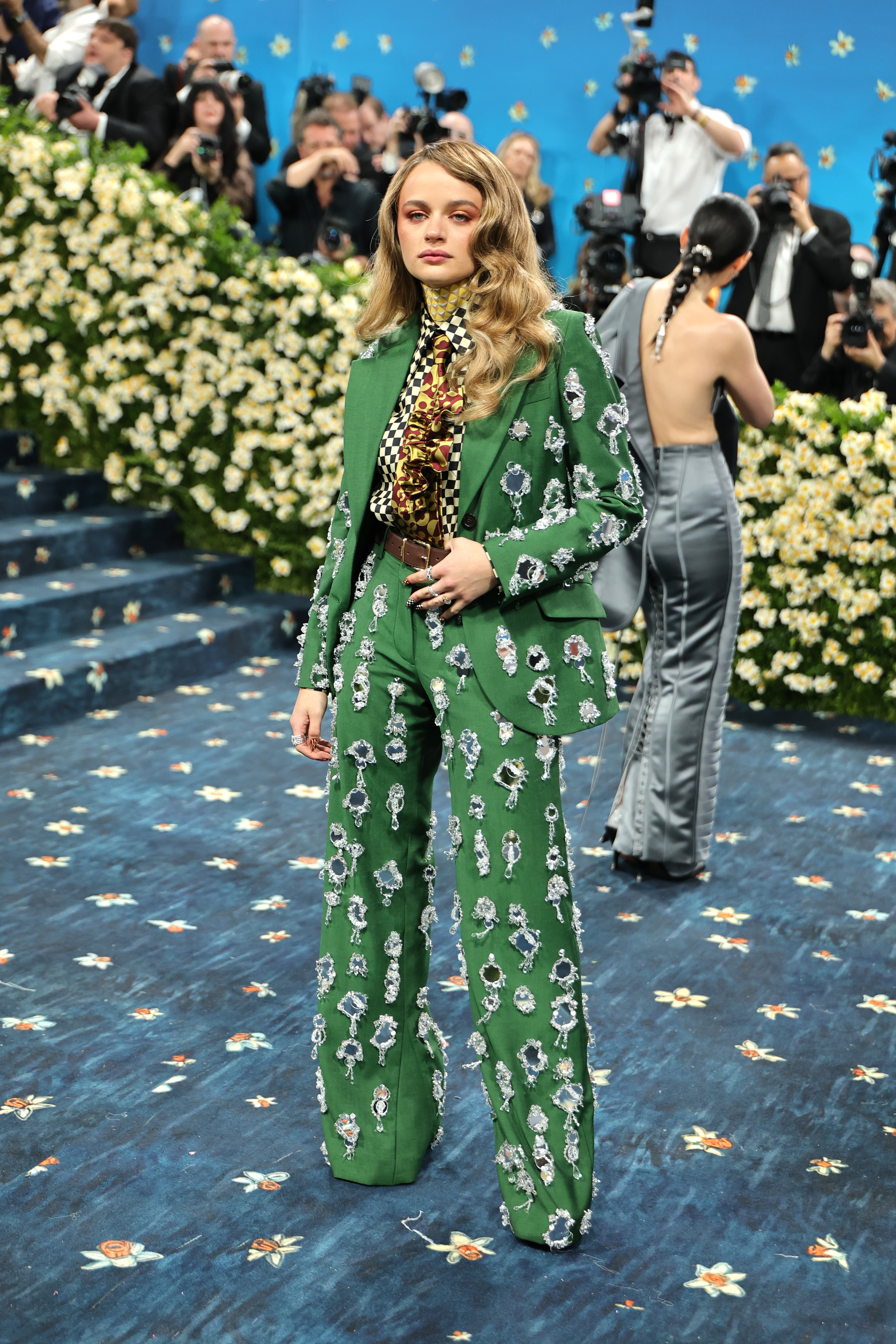 Person in patterned green suit poses on floral-themed carpet at event, surrounded by photographers and attendees