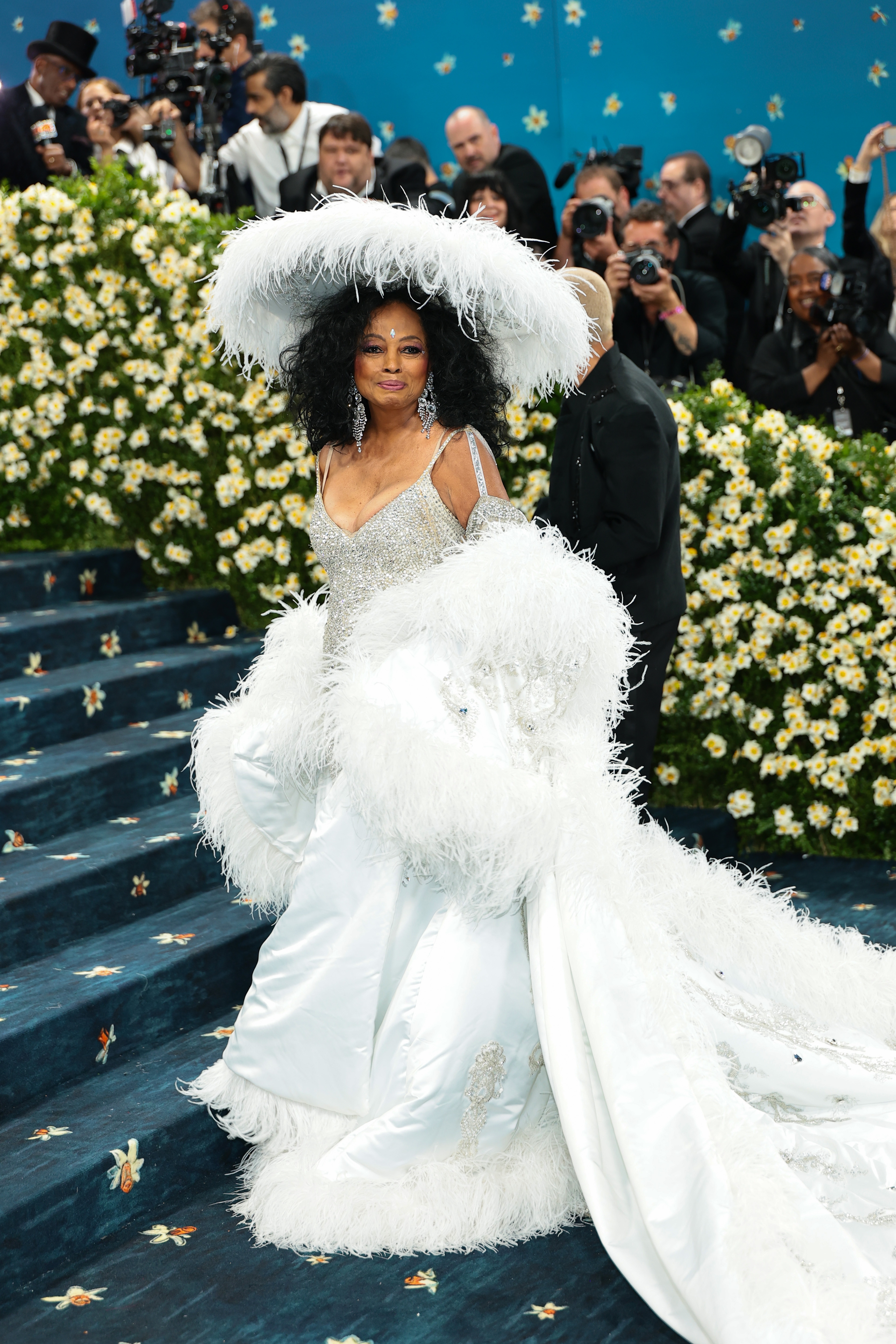 Person in a glamorous white feathered gown and wide-brimmed hat on a floral staircase at a formal celebrity event