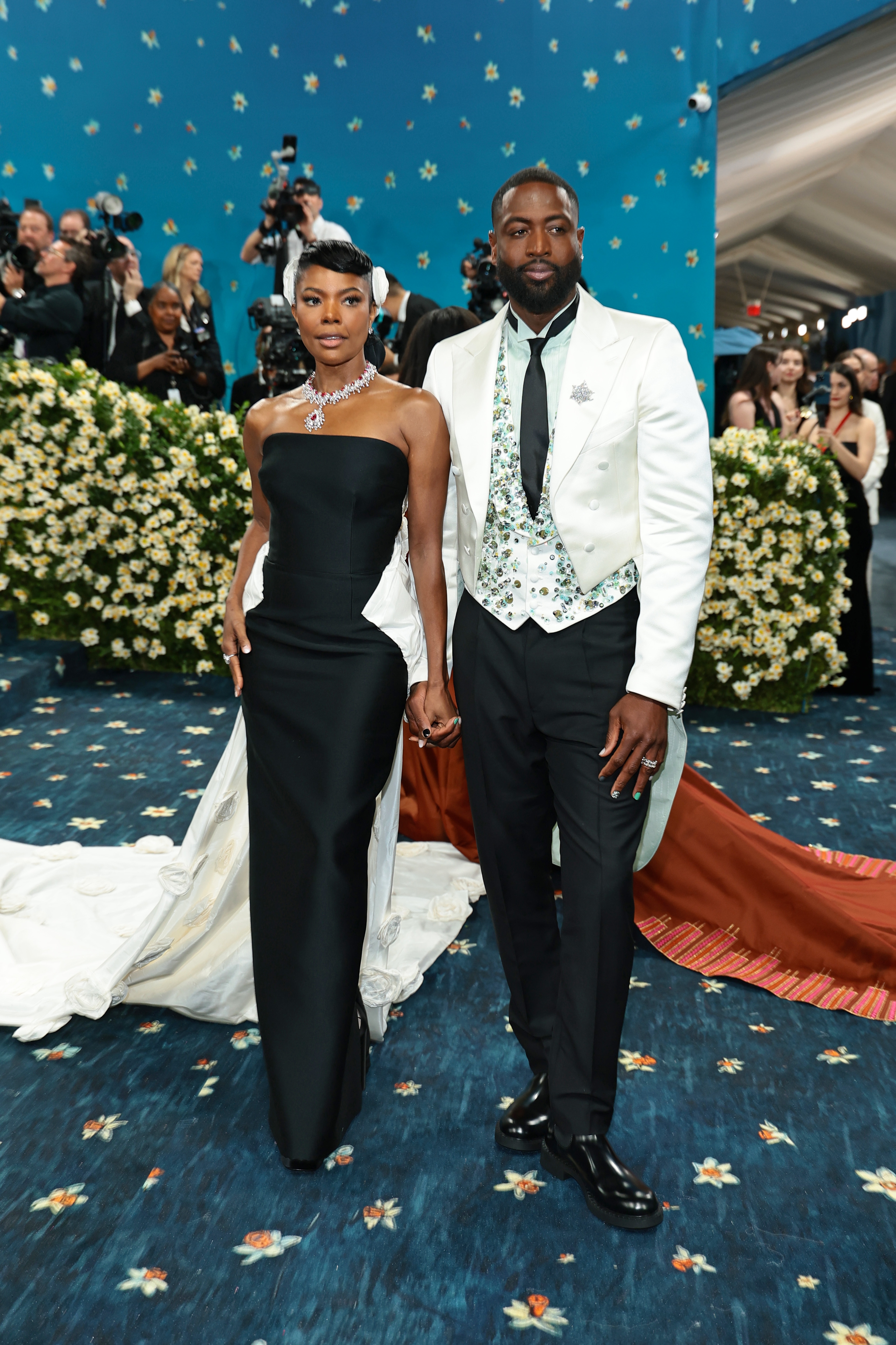 A couple on a red carpet, she in a strapless black gown with white train, he in a white tuxedo jacket with a patterned vest; holding hands