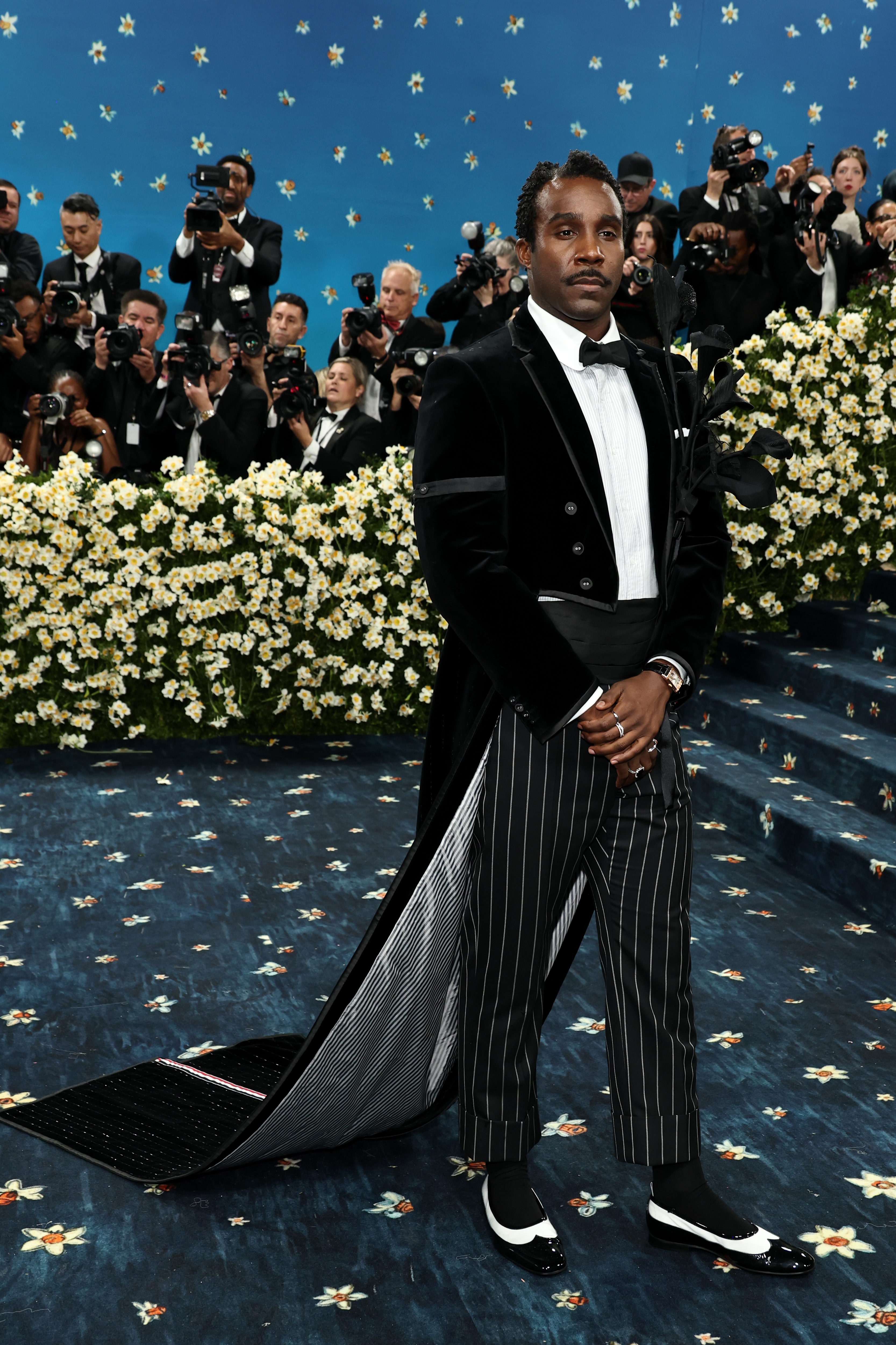 Person in a velvet tuxedo jacket with a long train and pinstripe pants poses on a starry-themed red carpet while photographers capture the moment