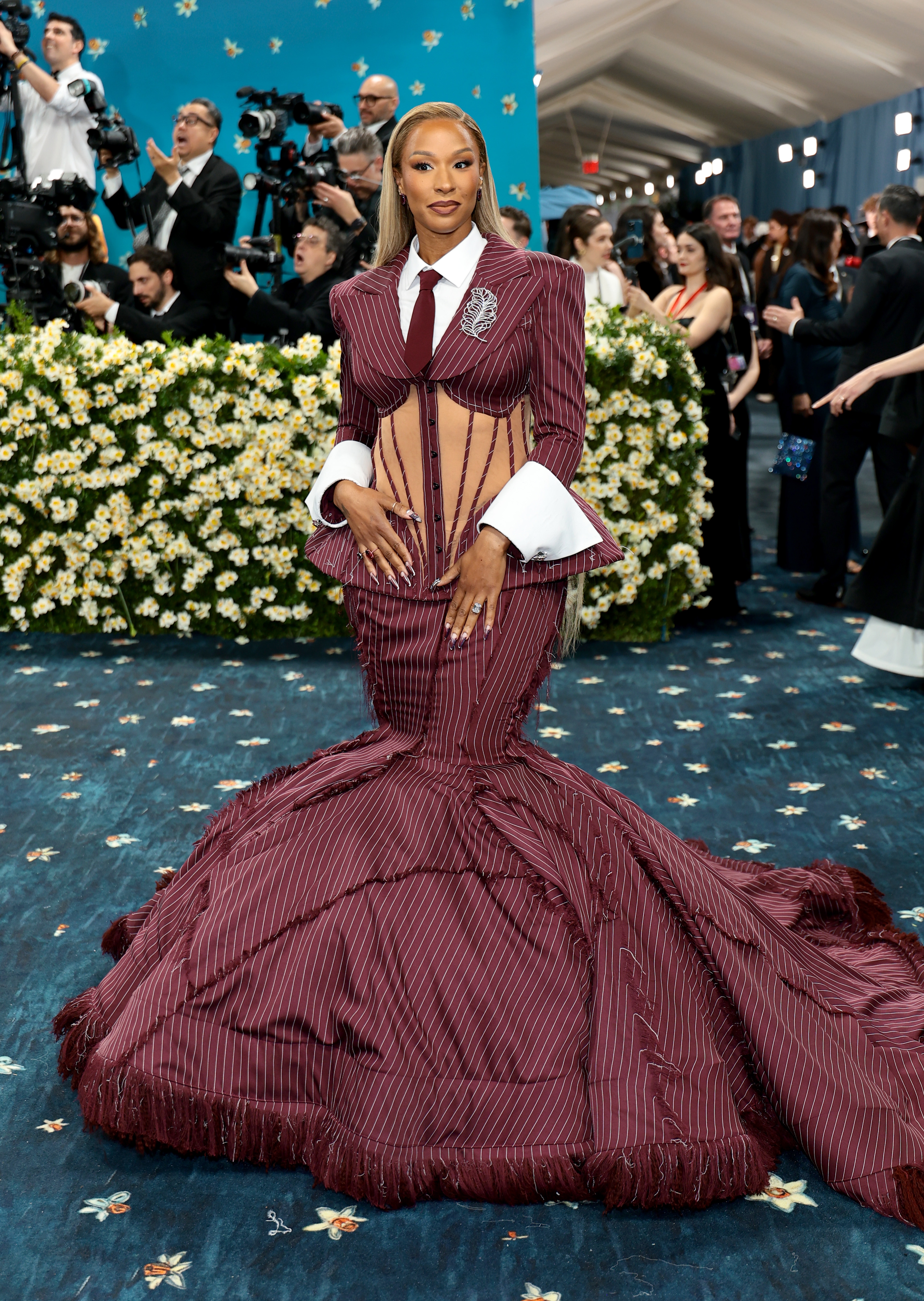 Person in a fitted, pinstripe gown with exaggerated shoulders and corset details on red carpet, surrounded by photographers and floral backdrop