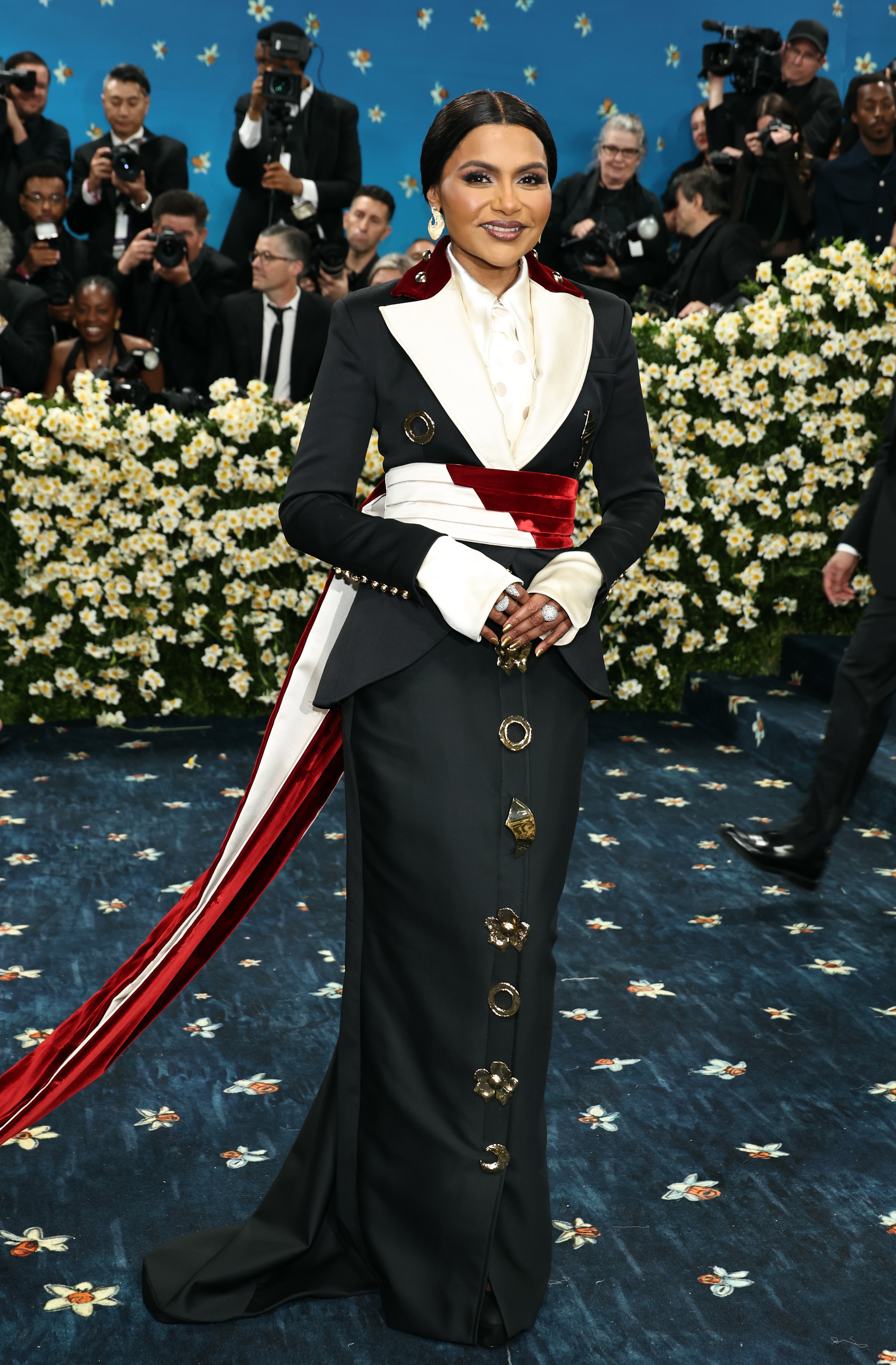 Woman in elegant black and white gown with red sash on a floral-themed red carpet with photographers in background