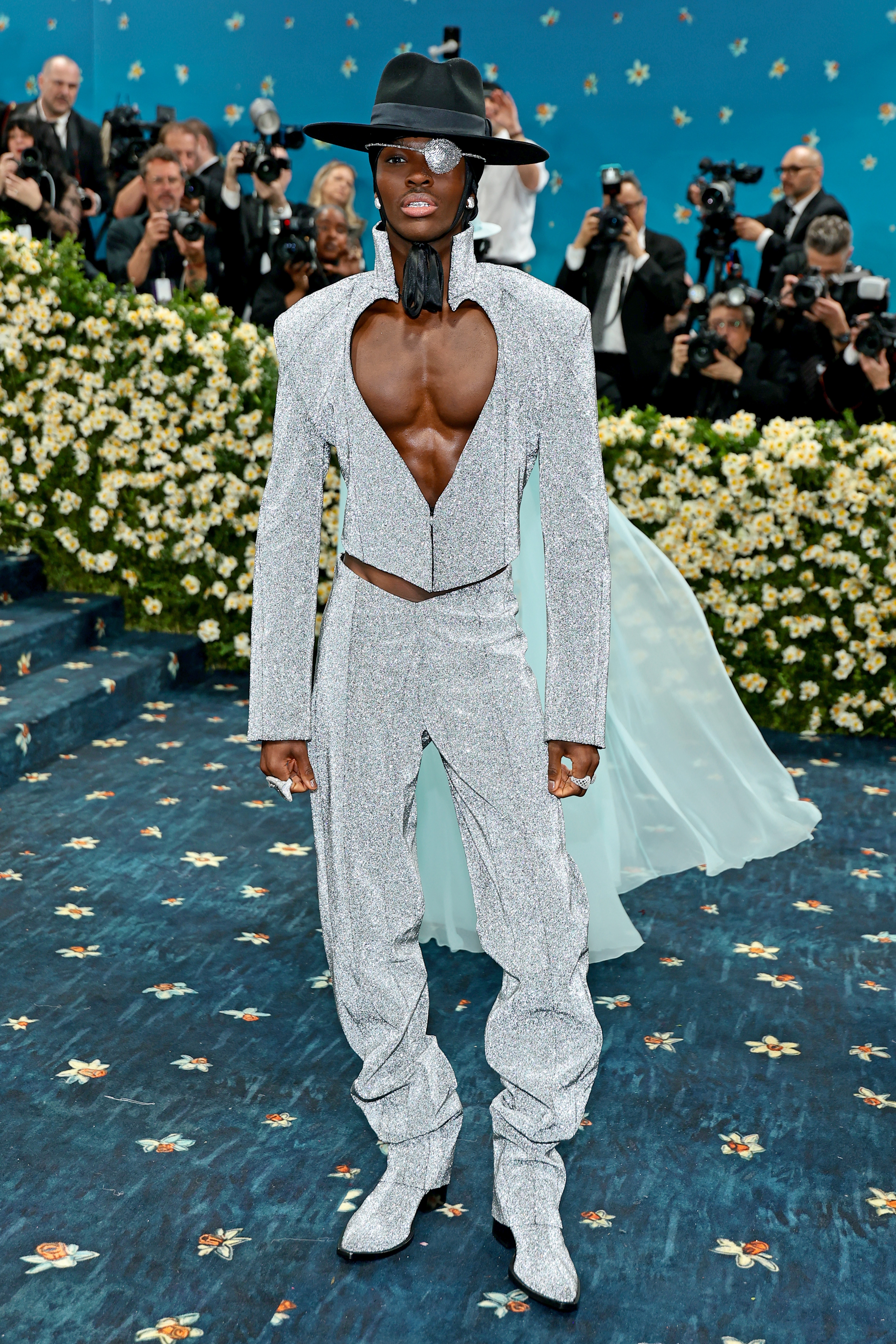 Person in a striking outfit with a deep V-neck, matching hat, surrounded by photographers at a celebrity event
