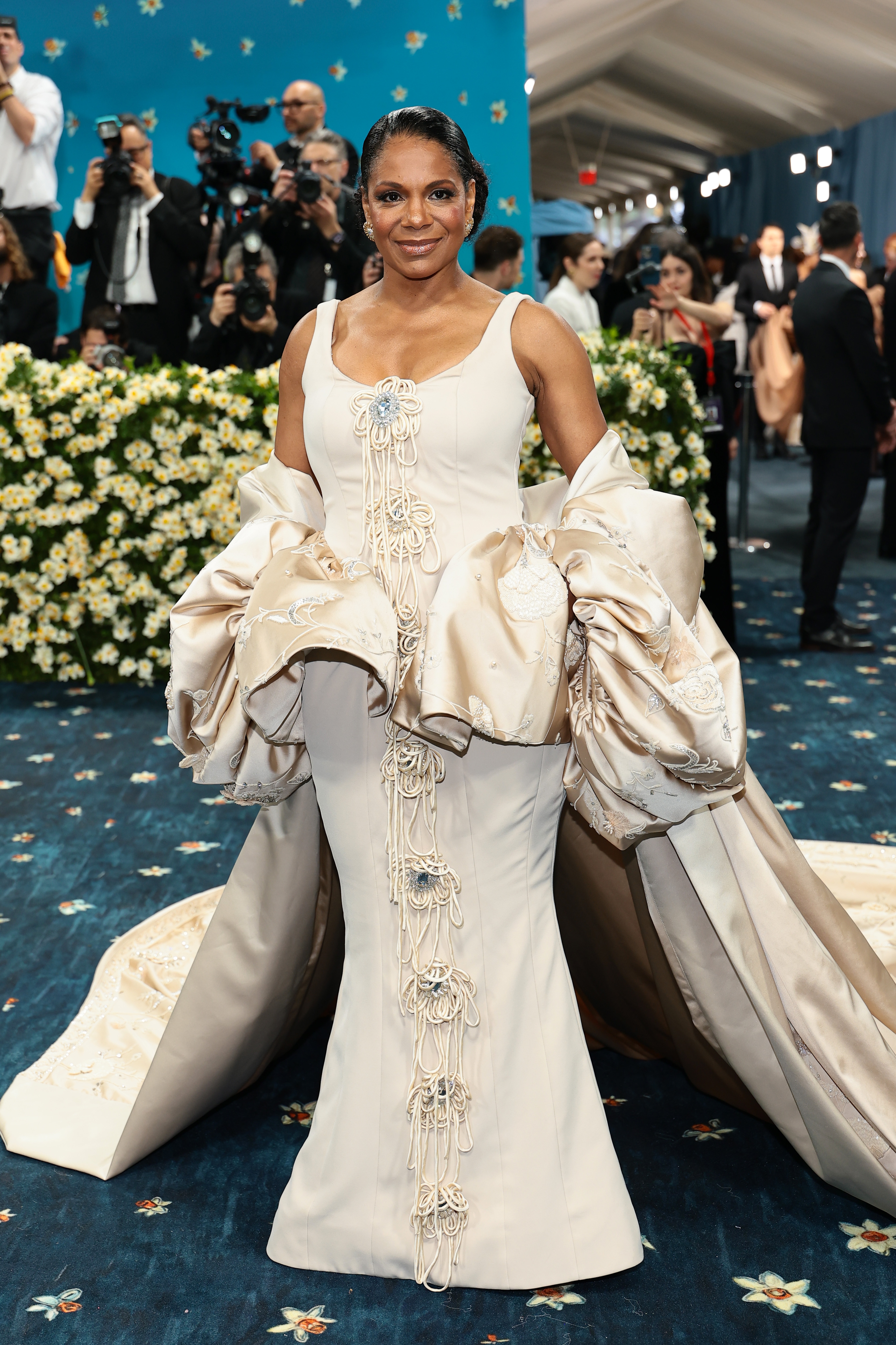 A person poses on a floral-patterned carpet wearing an elegant cream gown with elaborate drapery and intricate detailing, surrounded by photographers
