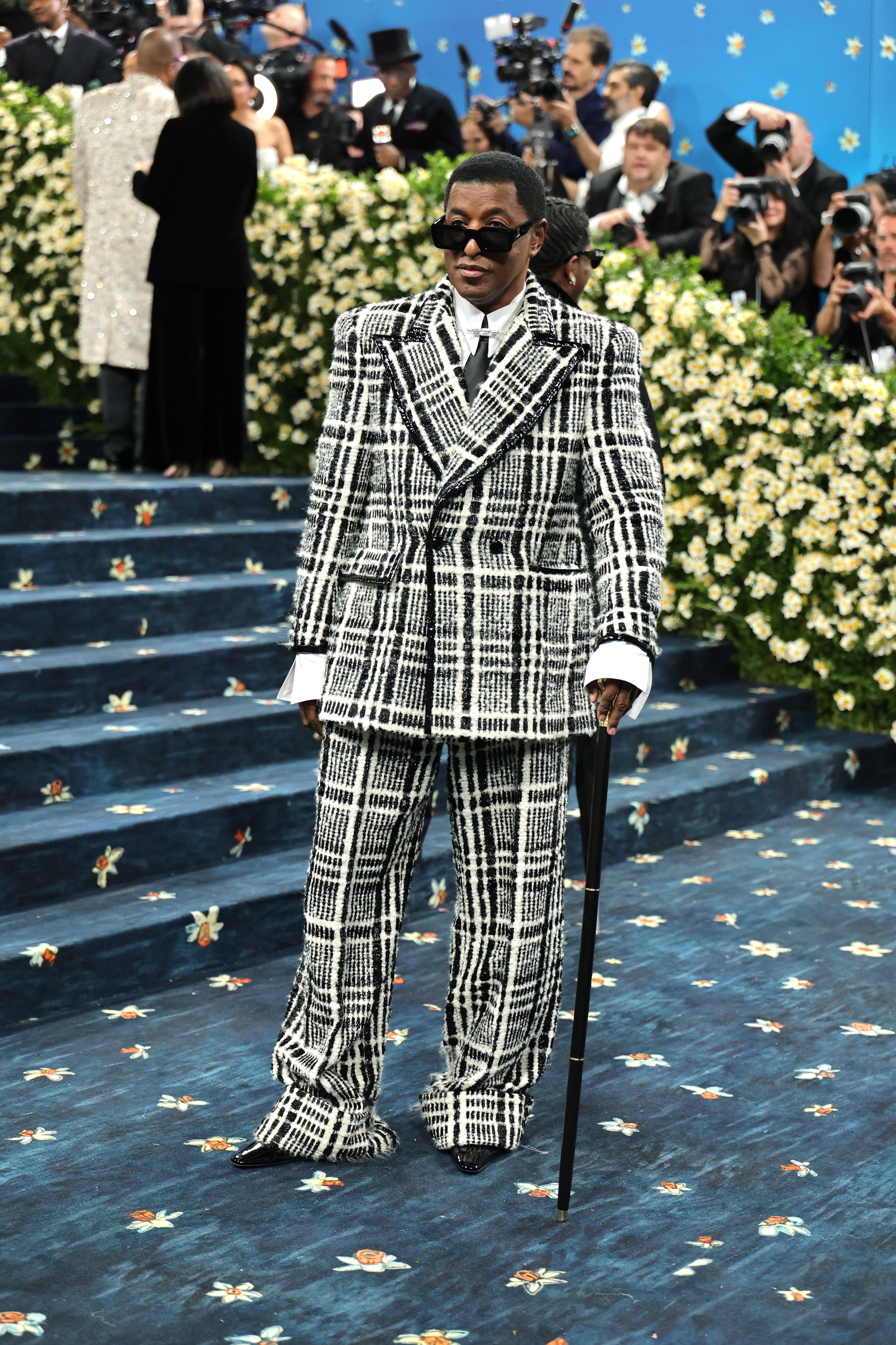 Person in a plaid suit with sunglasses and a cane on a blue floral pattern carpet at a formal event with onlookers