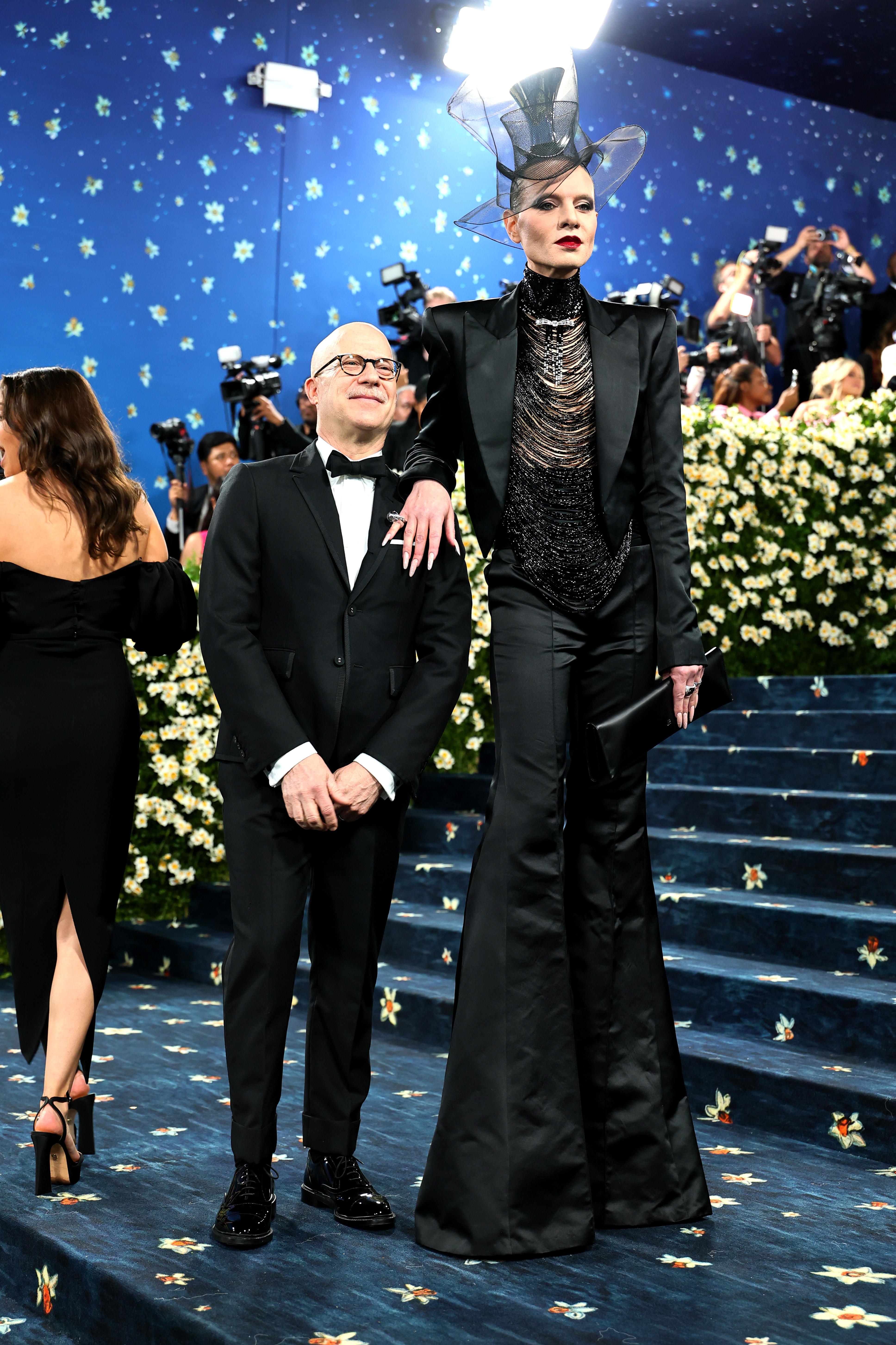 Two people pose on a floral staircase at a formal event. The taller person wears an avant-garde hat and black suit with sequined details