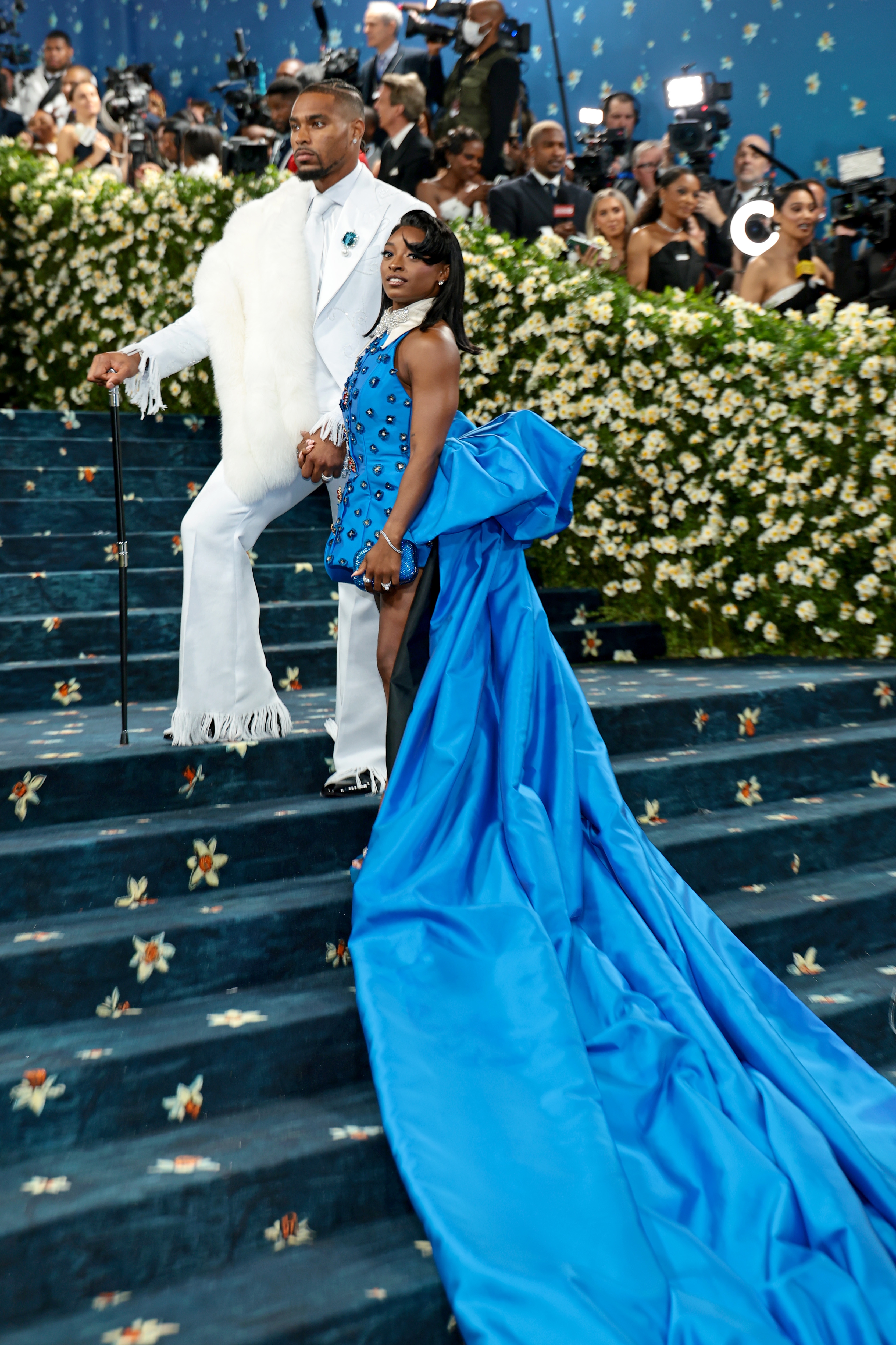 Two people on a red carpet. One wears a white suit with a cape and cane, the other a short dress with a long blue train