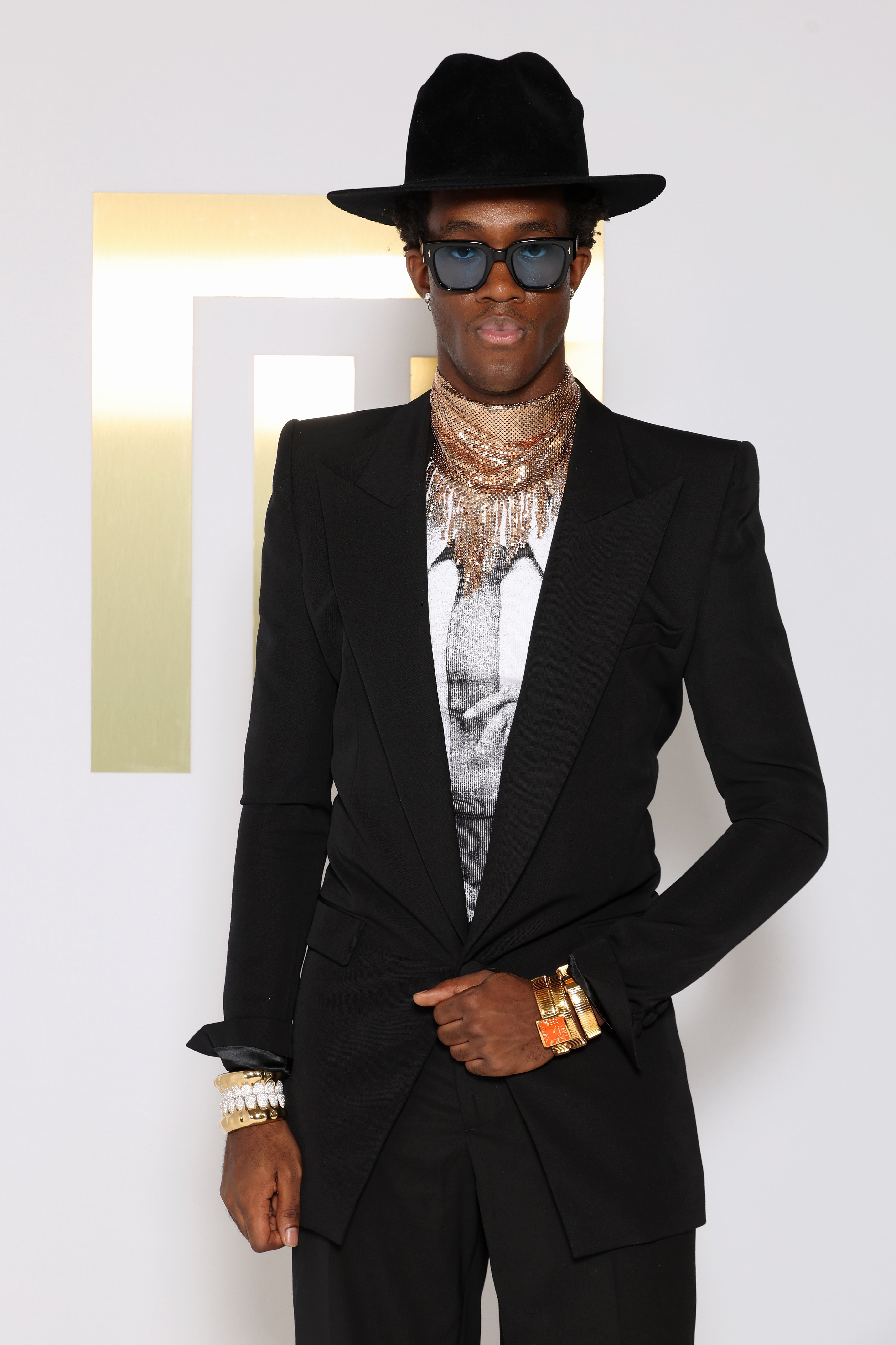 Person in a black suit with gold accessories, wide-brimmed hat, sunglasses, and layered jewelry at a formal event
