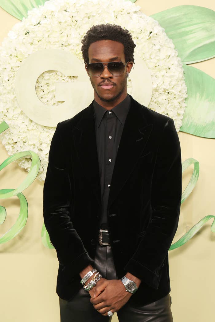 A man in a velvet suit and sunglasses poses on a red carpet with a floral backdrop at a celebrity event