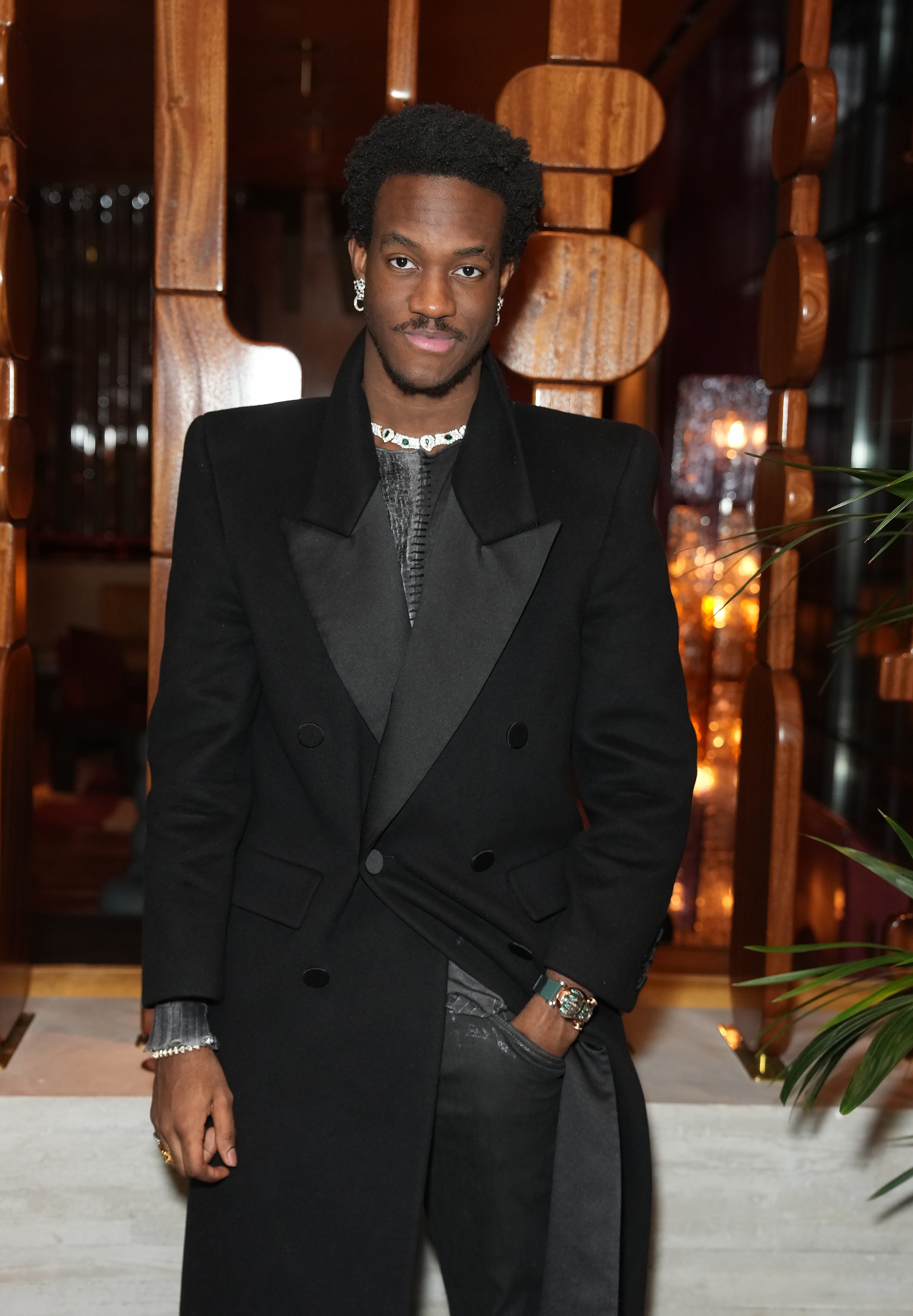 Person in a stylish long black coat with satin lapels, wearing a chain necklace and earrings, attending an elegant event