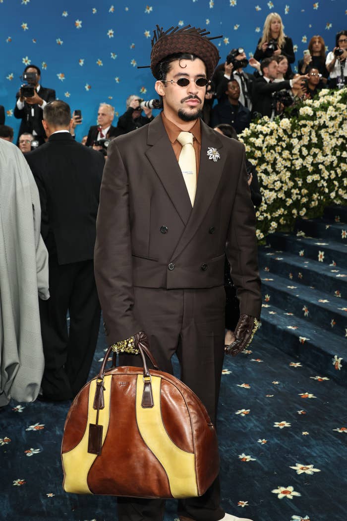 Persona con traje formal de dos piezas, corbata clara, guantes ornamentados y bolso grande, posando en un evento glamuroso con fondo decorado